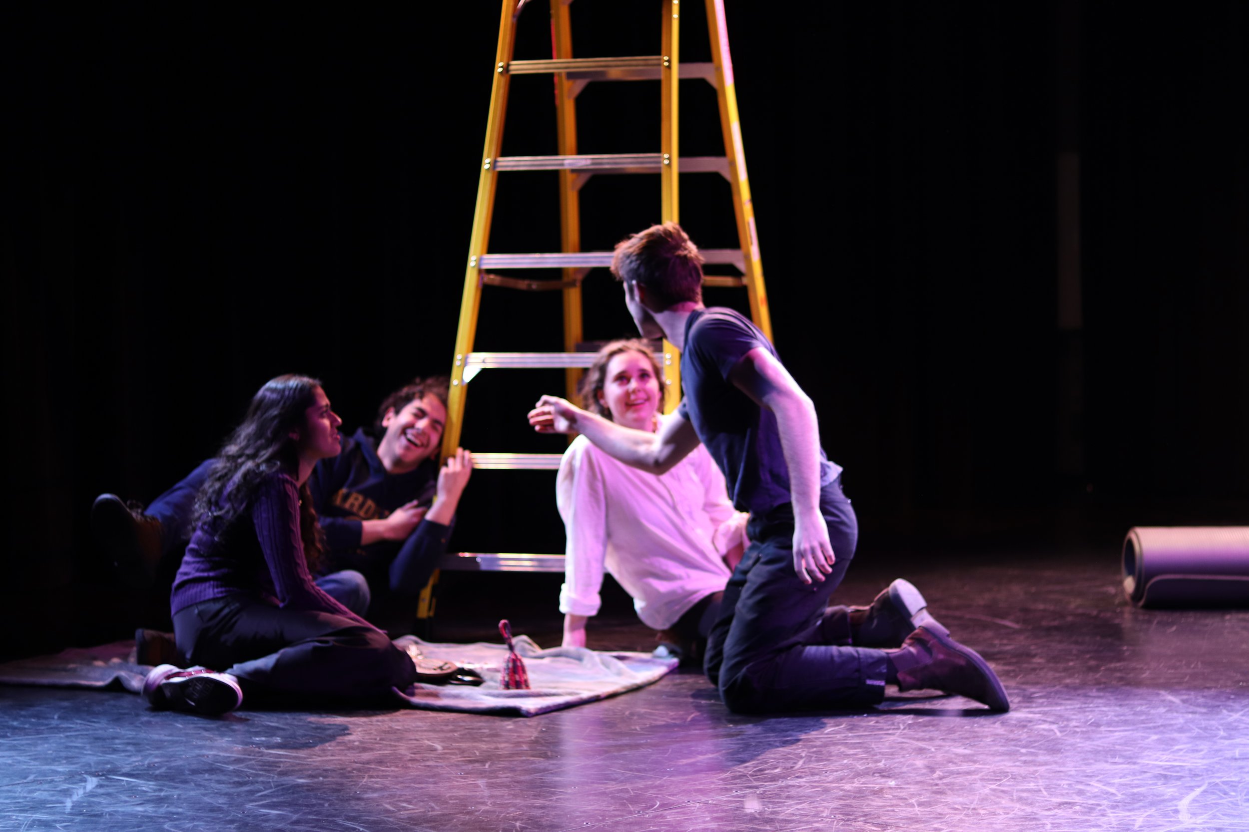 A group of five young people on stage with a ladder in the background, sitting and kneeling around a blanket, engaged in a theatrical scene, with one person standing and speaking animatedly.