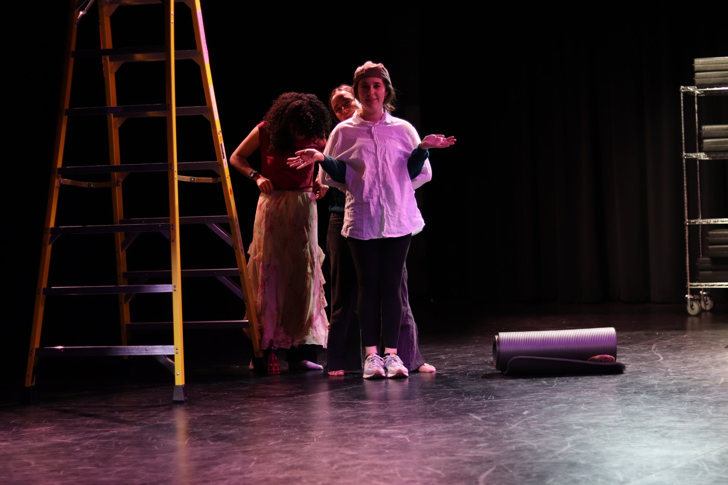 Four children stand on a stage, with a yellow ladder on the left and a rolling cart on the right. The children are in casual clothing, appearing to be rehearsing or performing.