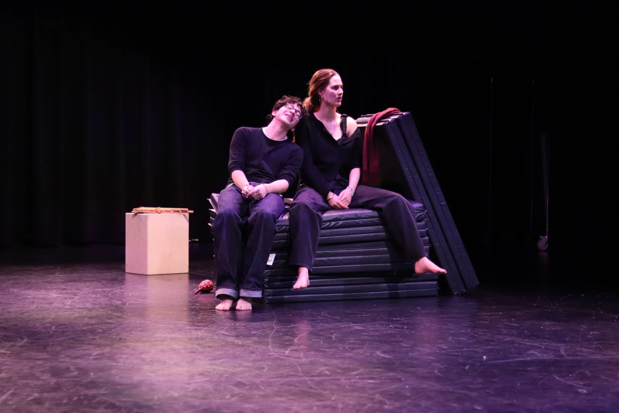 Two women sitting on a black stage set, one resting her head on the other's shoulder, with props including a small wooden box and a black padded platform.