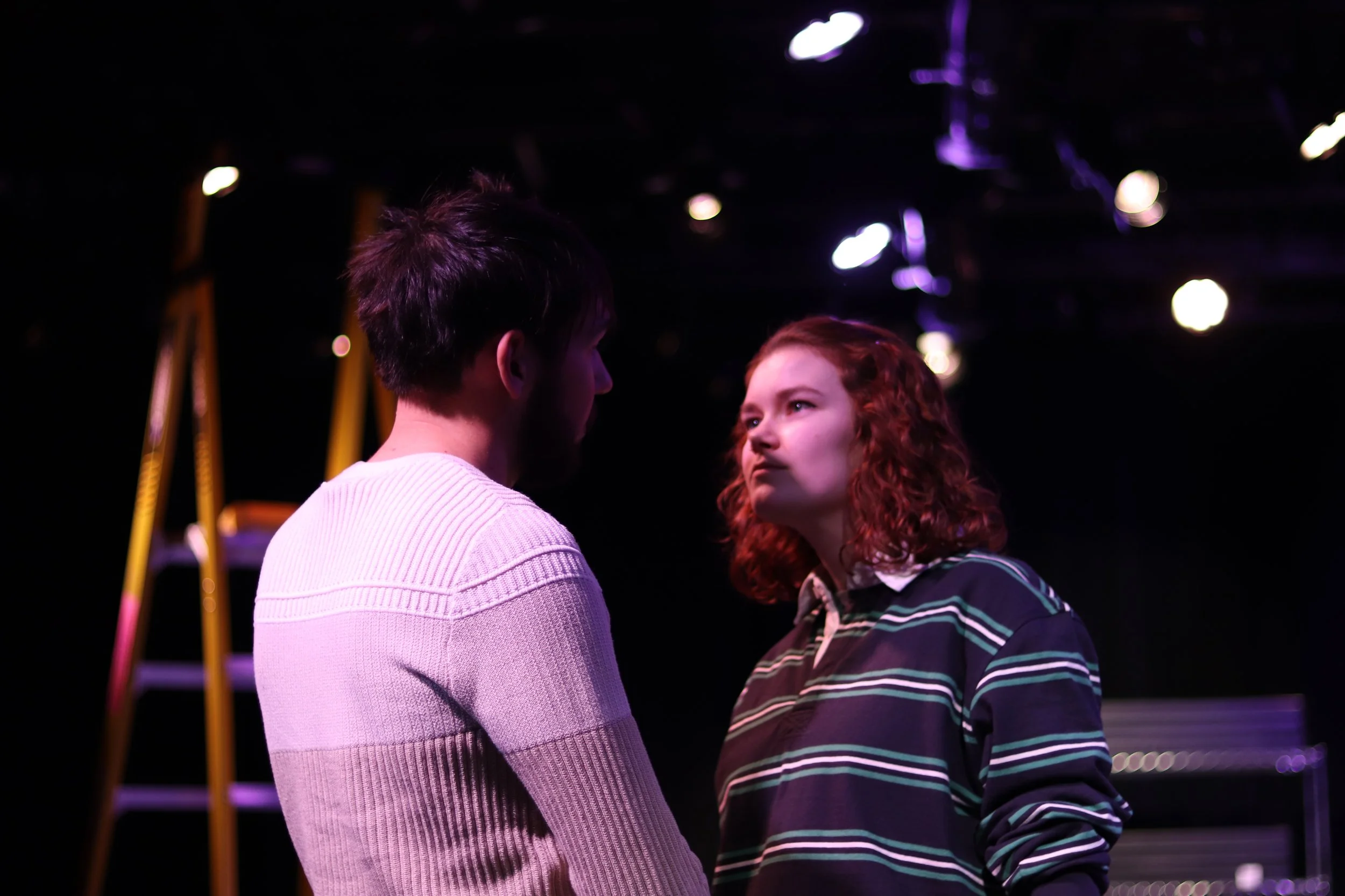 Two young people, a man and a woman, face each other in a dark room with stage lighting, with a ladder and stage equipment in the background.