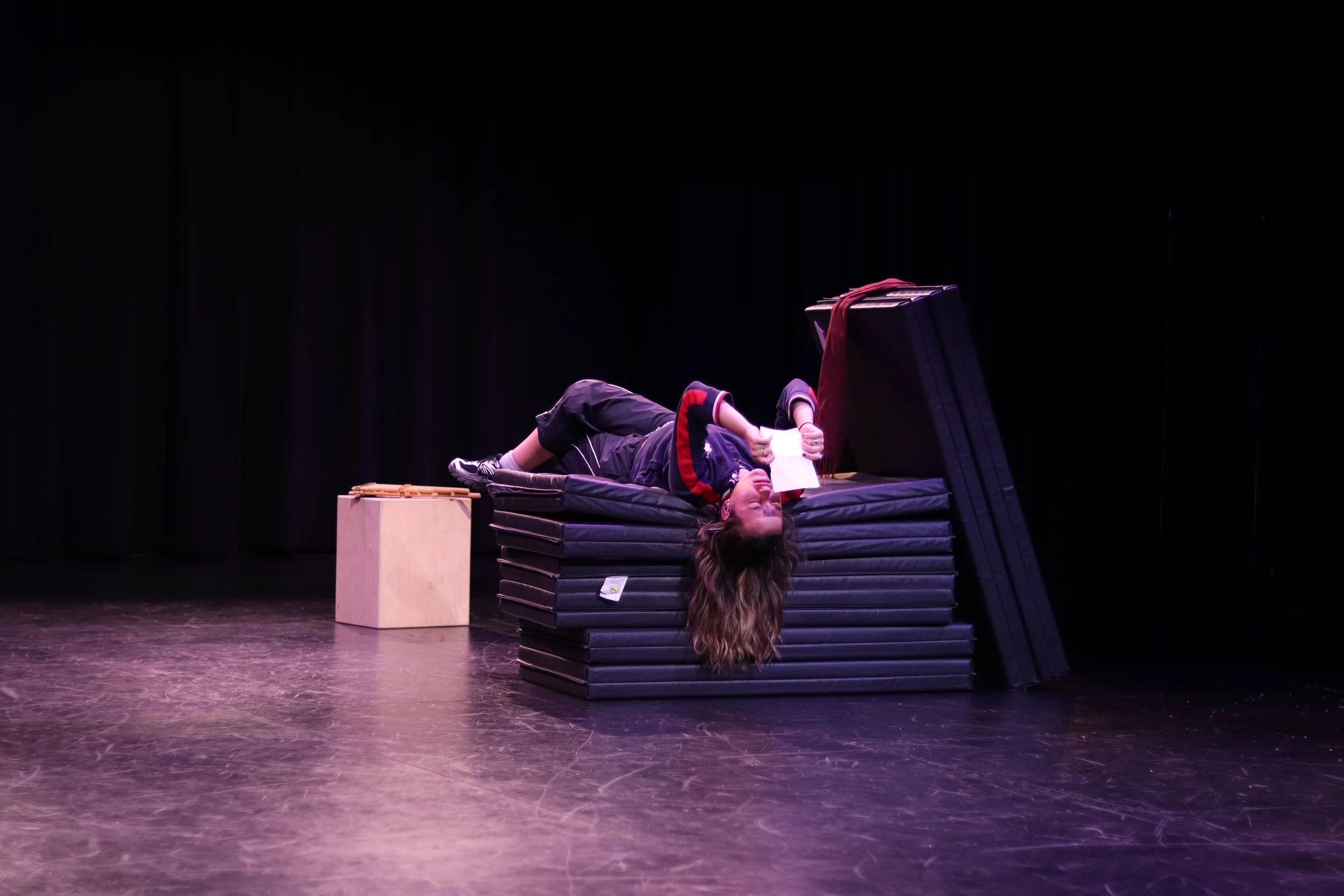 A woman lying on a stack of black gym mats on a stage, holding a sheet of paper, with a small wooden cube nearby and a large black case leaning against the mats