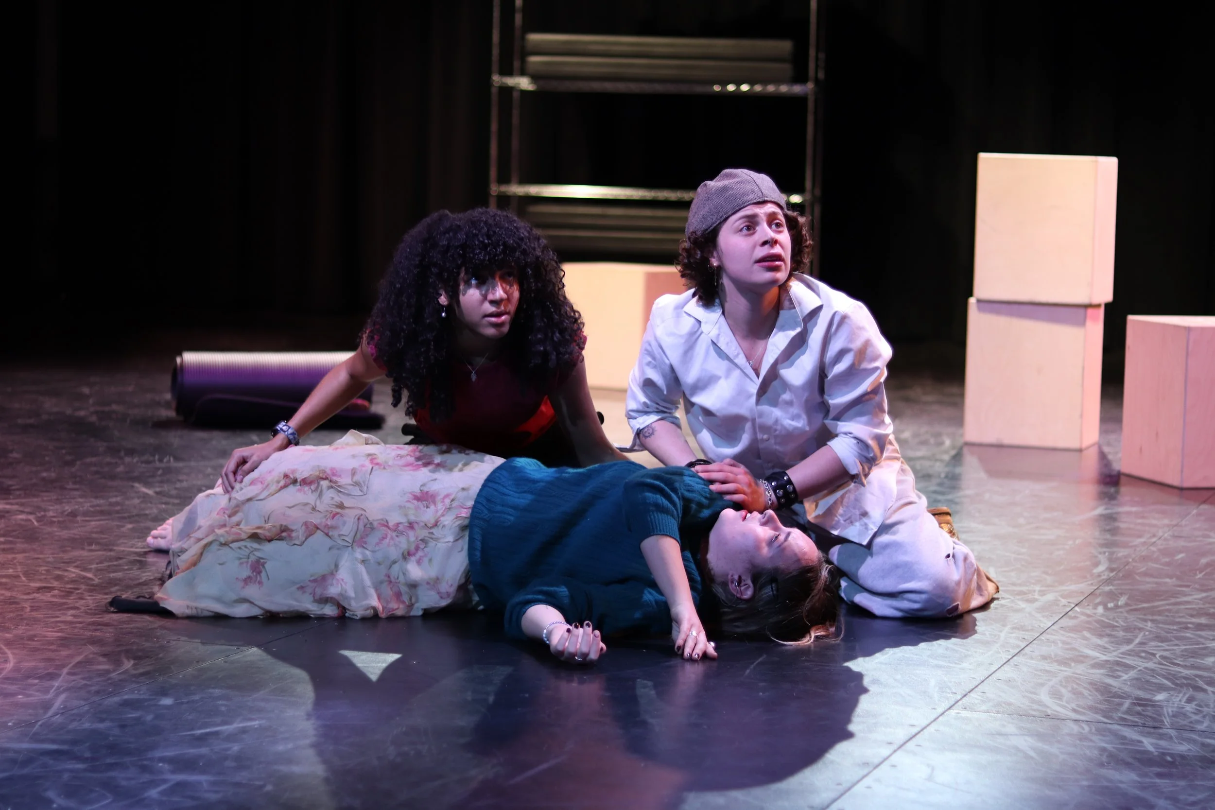 Three young women on stage, one lying on the floor and the other two kneeling beside her, with dramatic expressions, in a theatrical performance.