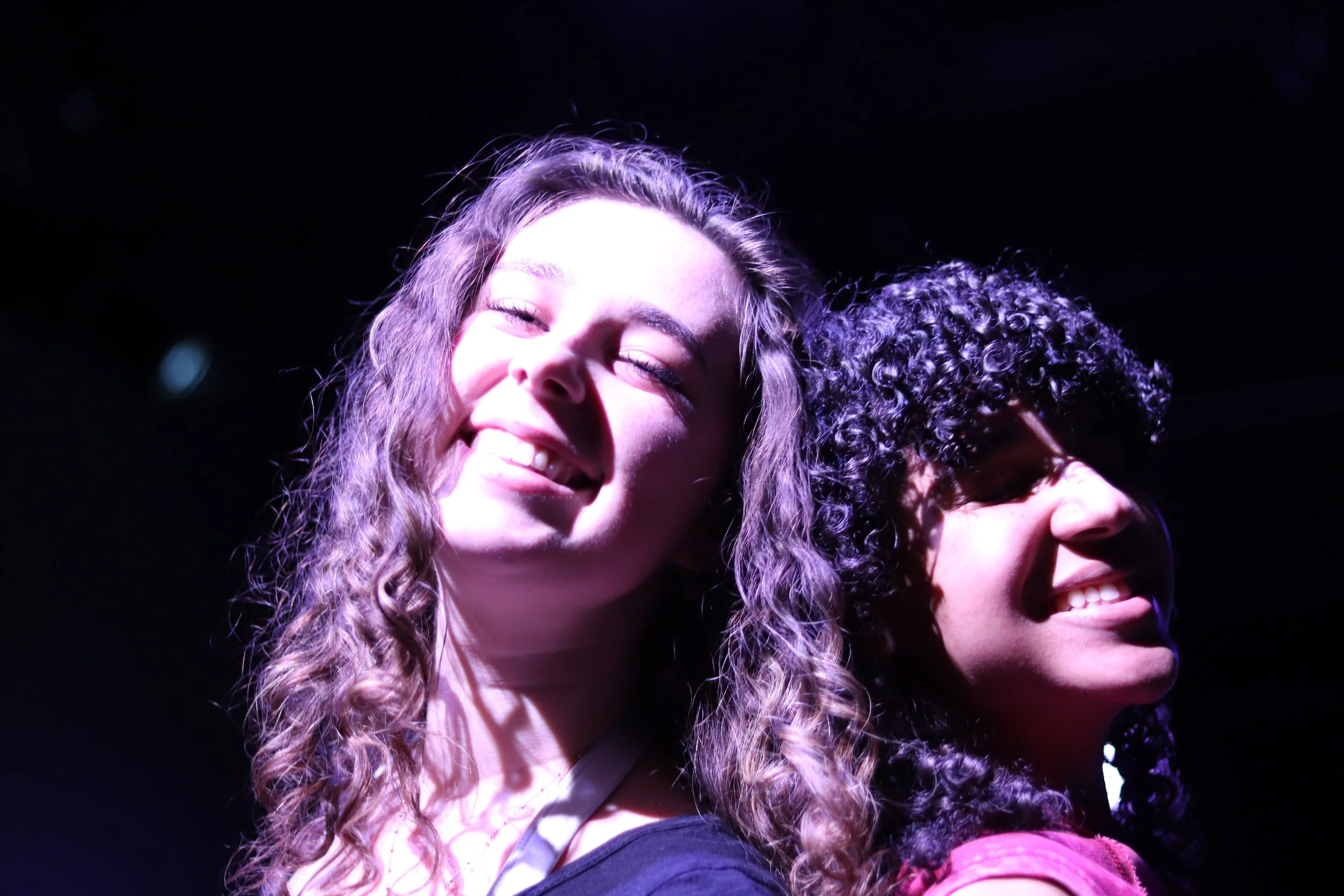 Two smiling young women with curly hair standing back-to-back in bright purple lighting, facing the camera.