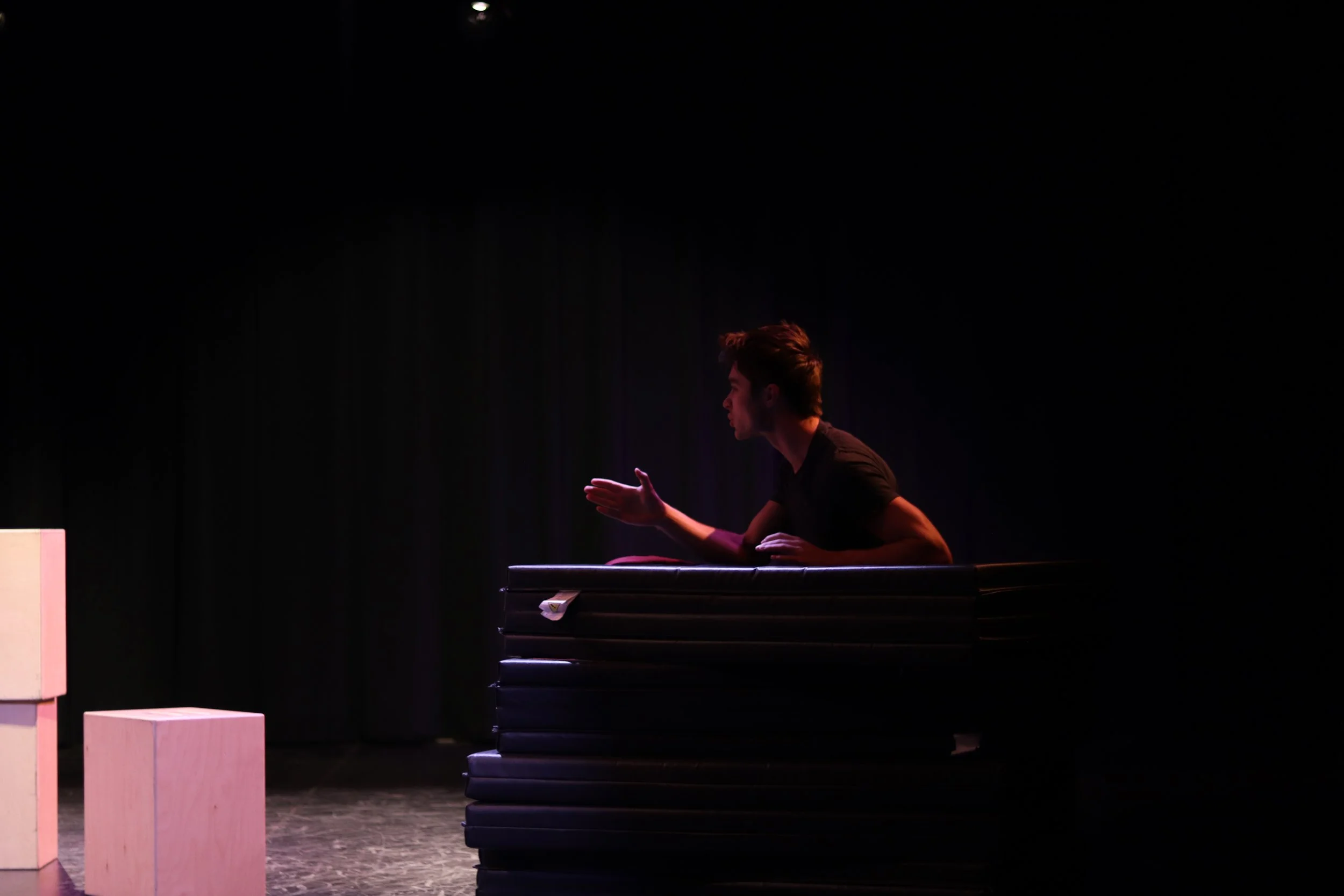 A young man performing on stage with a dark background, gesturing with his hand while leaning on a platform.