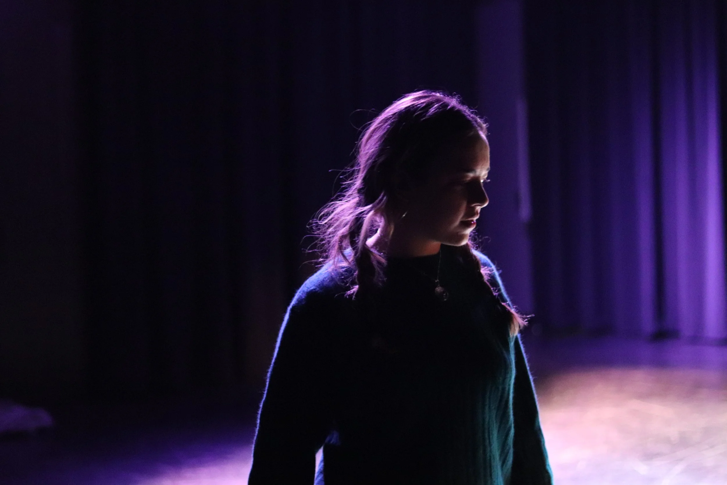 A young woman with braided hair stands on stage, illuminated by purple stage lights, with a dark curtain in the background.