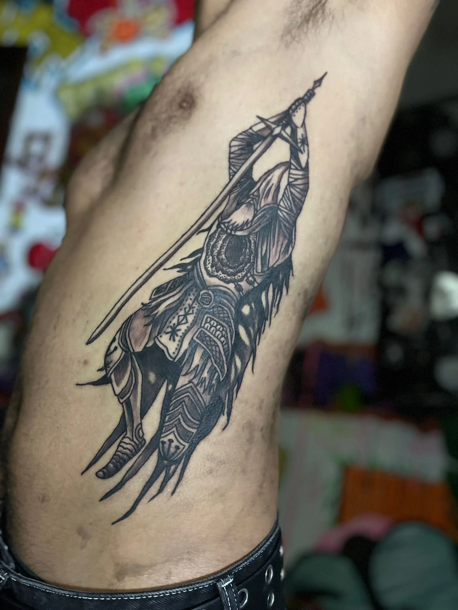 Black and gray tattoo of a warrior's head with armor and a feathered headdress, holding a spear, on a person's side ribcage.