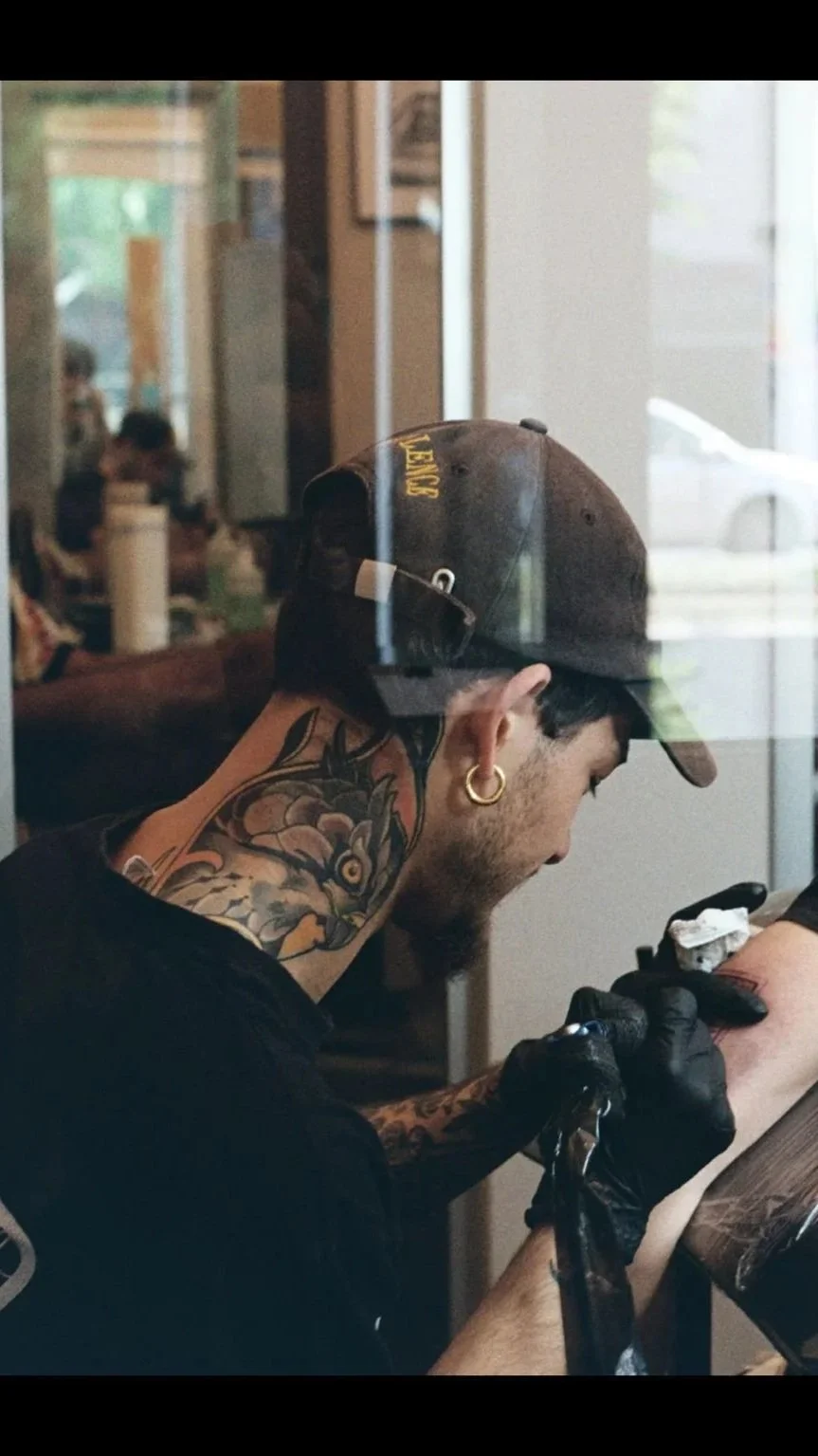 A man with tattoos on his neck and arms, wearing a cap with yellow lettering, is tattooing another person's arm in a tattoo shop. The reflection of the scene can be seen on the window.