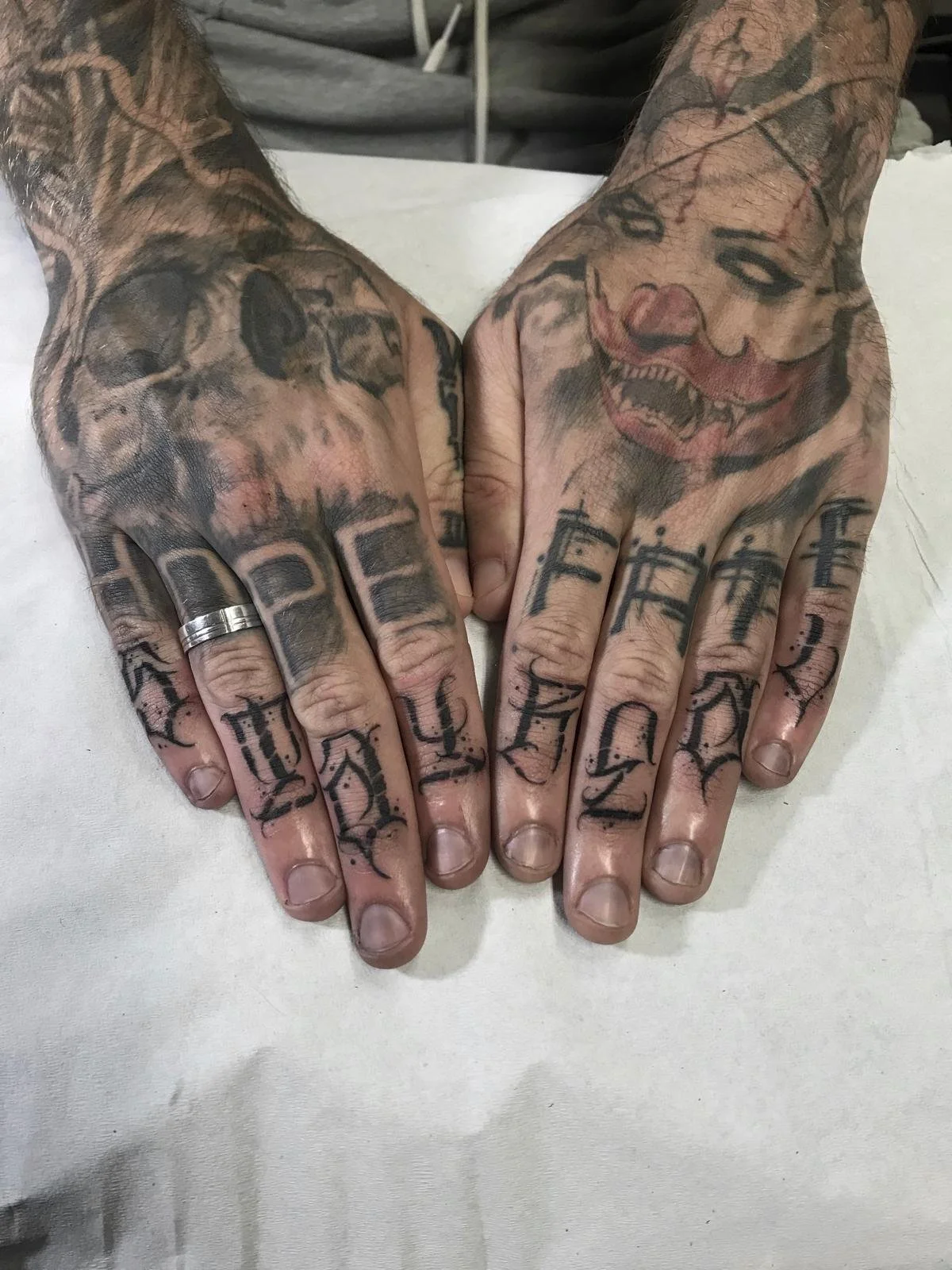 Two tattooed hands resting on a white surface, with the left hand showing black ink letters on the fingers and a skull tattoo on the back, and the right hand showing letters and a smiling clown face tattoo.