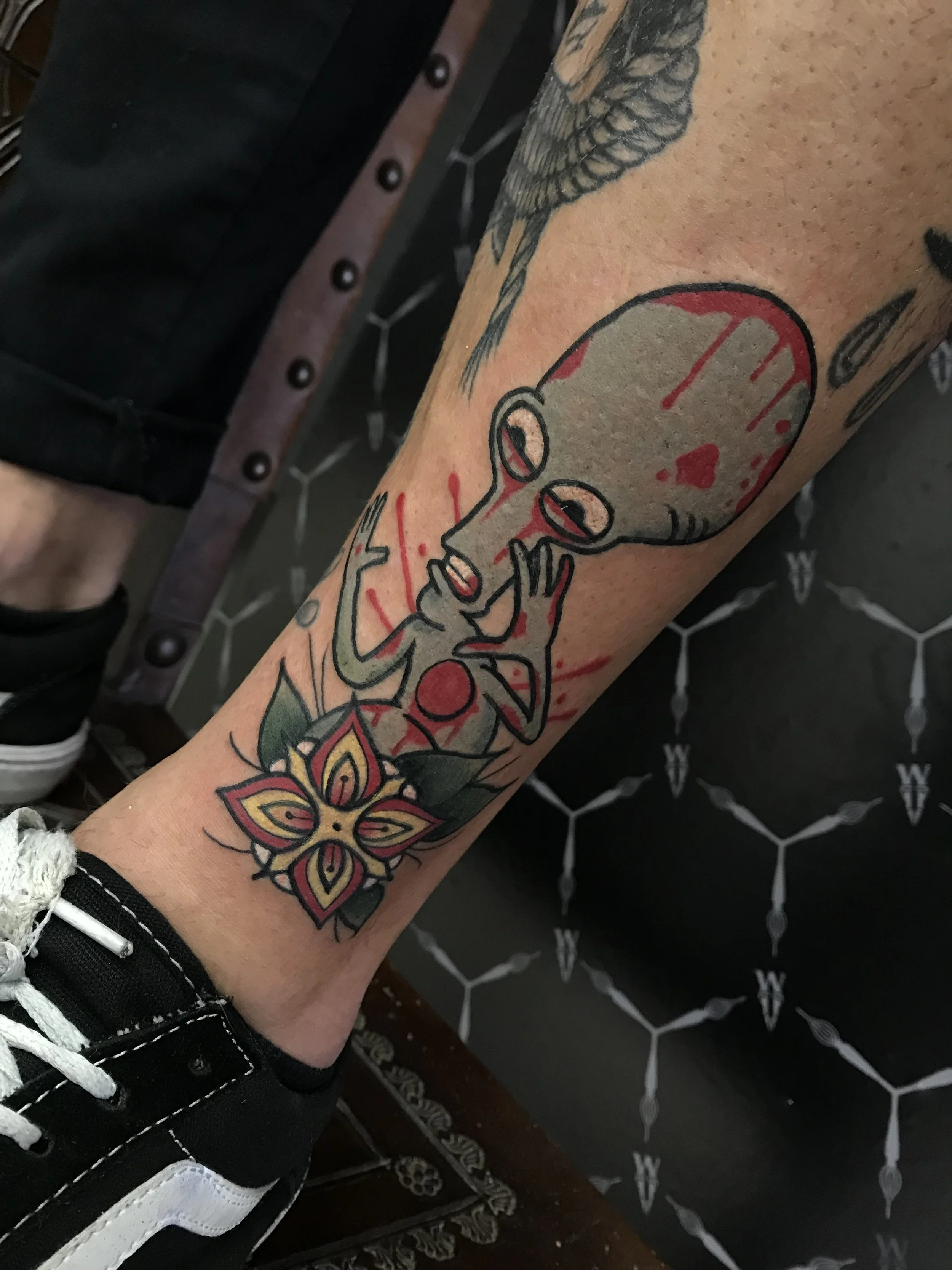 Tattoo of a large alien-like head with a small body, holding its face with both hands, surrounded by red splashes, with a floral design below.