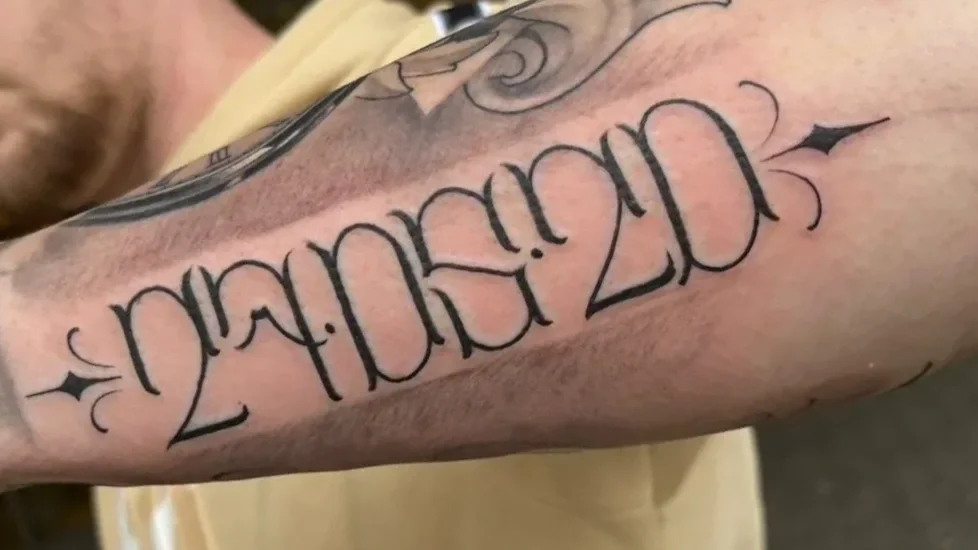 Tattoo on person's arm displaying the date 'January 20, 2024' in black ink with decorative elements.