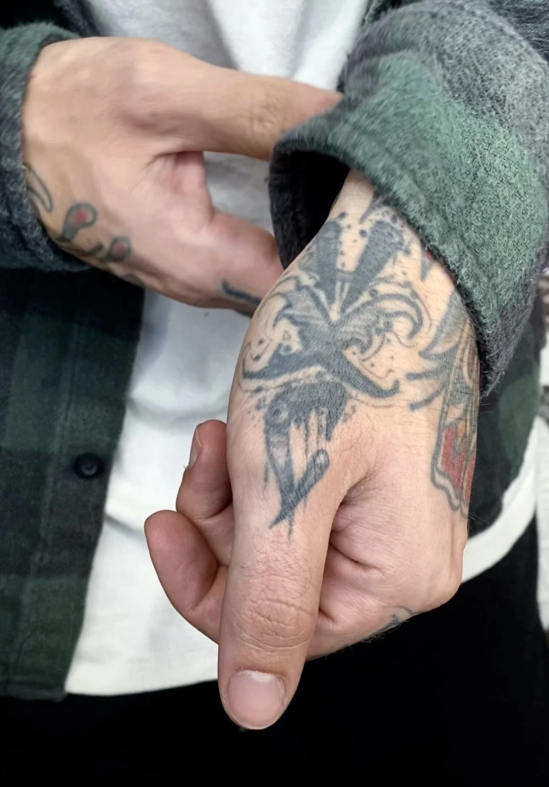 Close-up of a person's hand with tattoos, wearing a green and gray plaid sleeve, with the middle finger extended and the ring finger bent. Additional tattoo visible on the wrist.