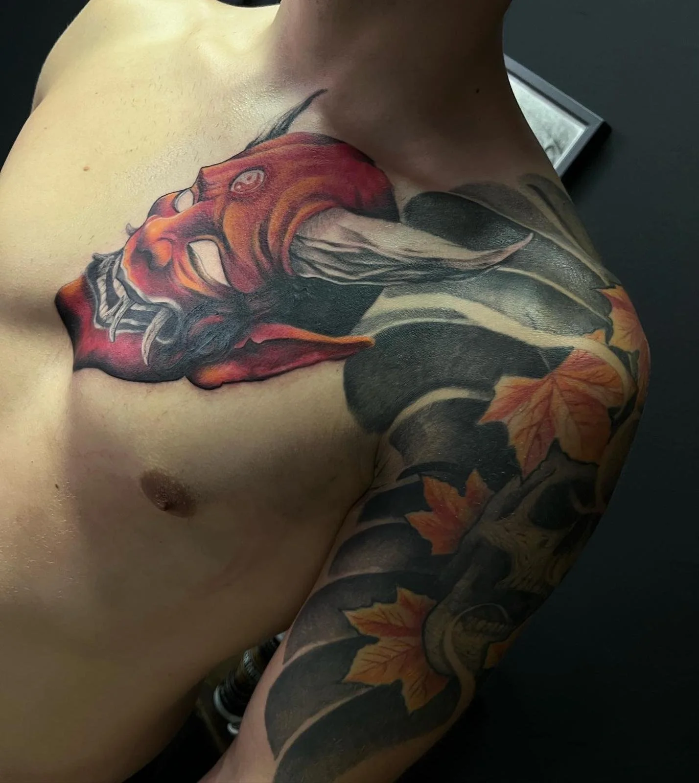 Tattoo of a red and black dragon head on a person's shoulder, with autumn leaves on the arm.