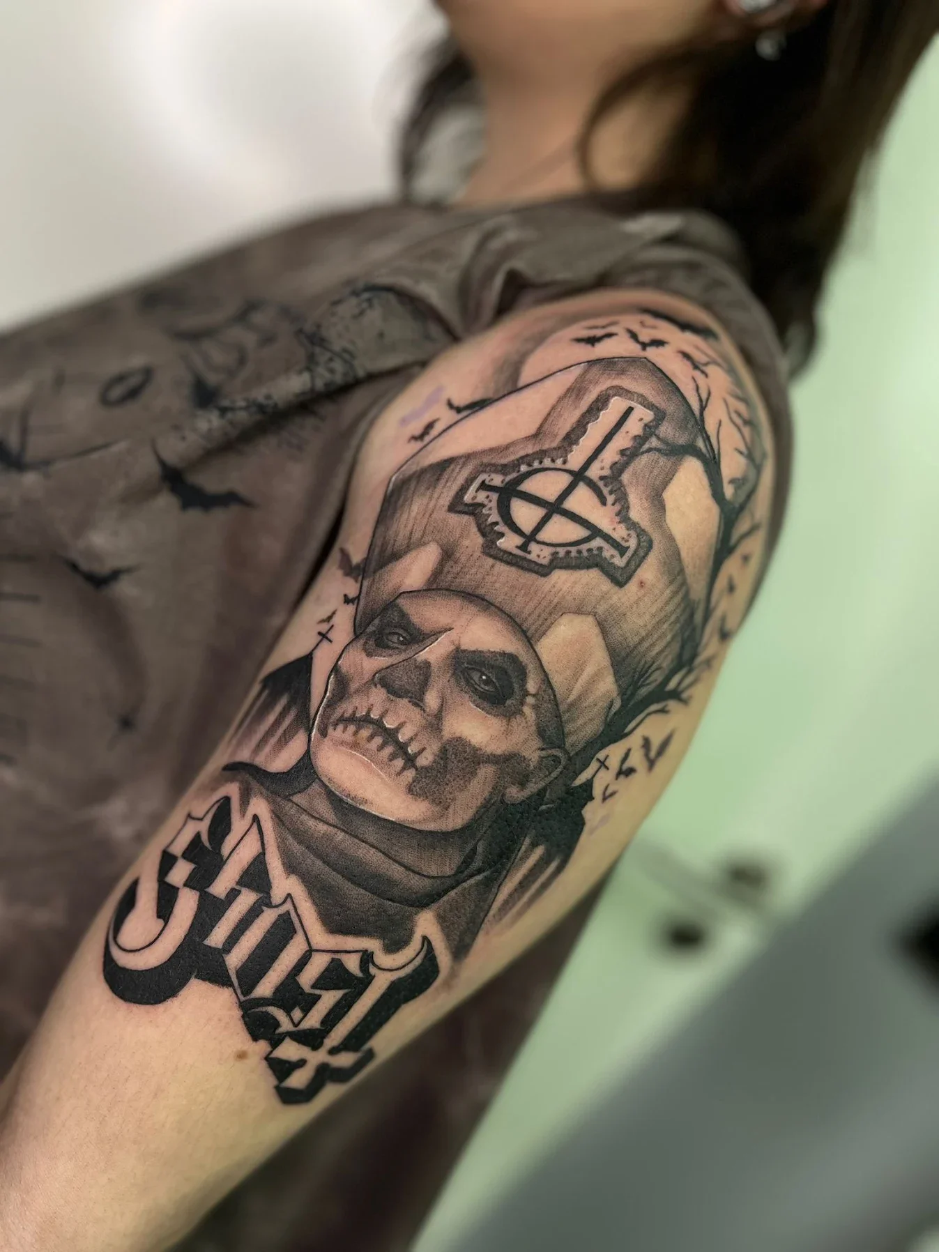 Black and gray tattoo of a skull with dark eye sockets and teeth, surrounded by various symbols, and the word 'death' at the bottom on a person's upper arm.