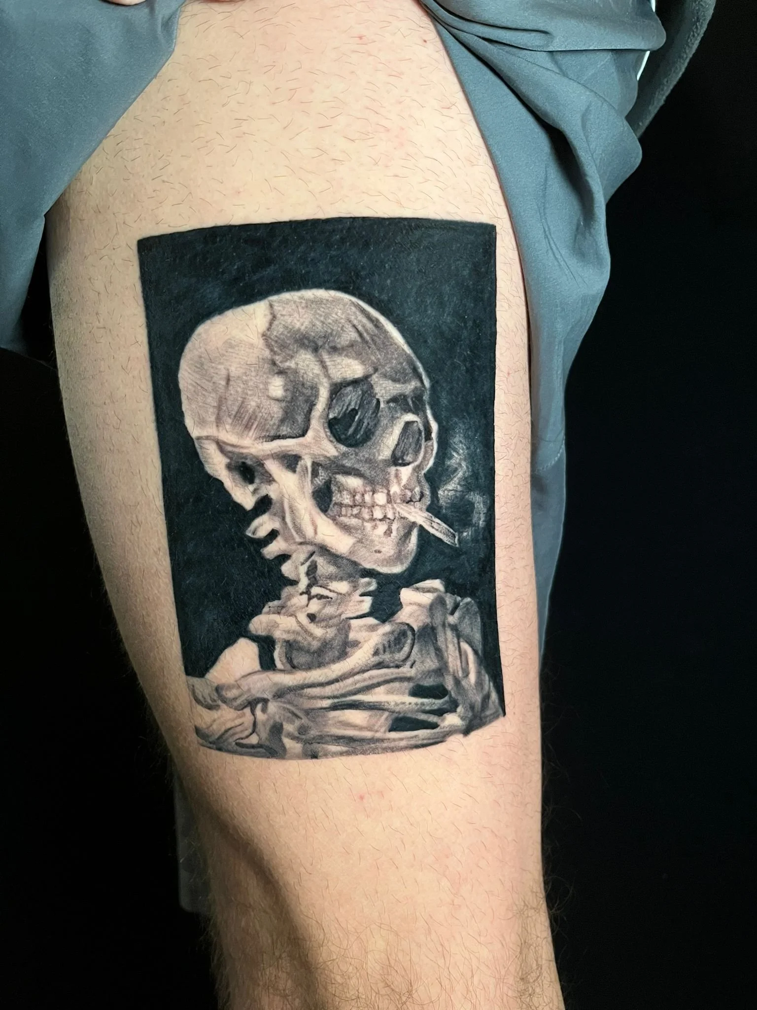 Black and gray tattoo of a human skull with a cigarette in its mouth, on the upper arm of a person.