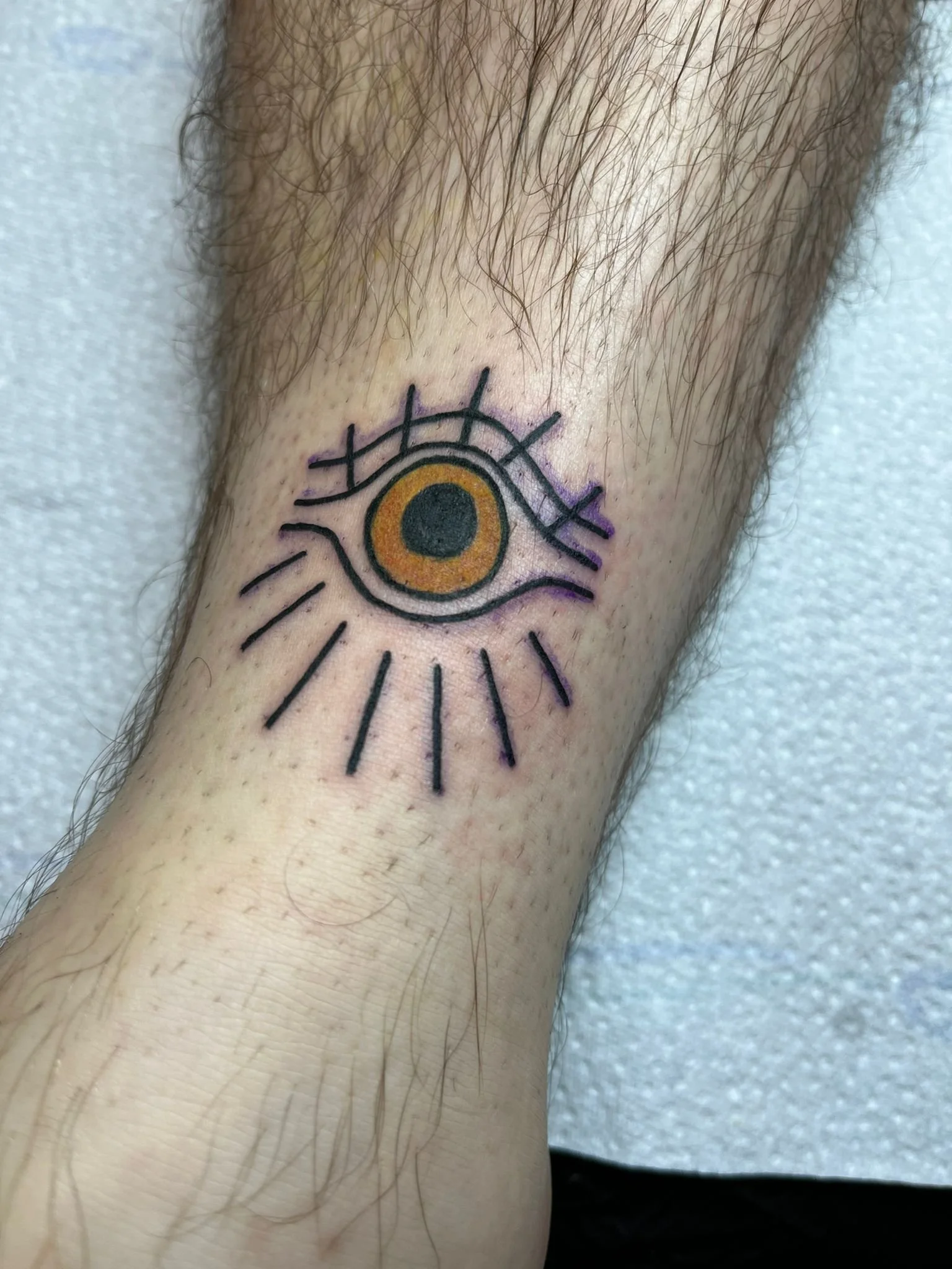 Tattoo of an eye with orange and black iris and long black lines radiating from below, on a person's arm.