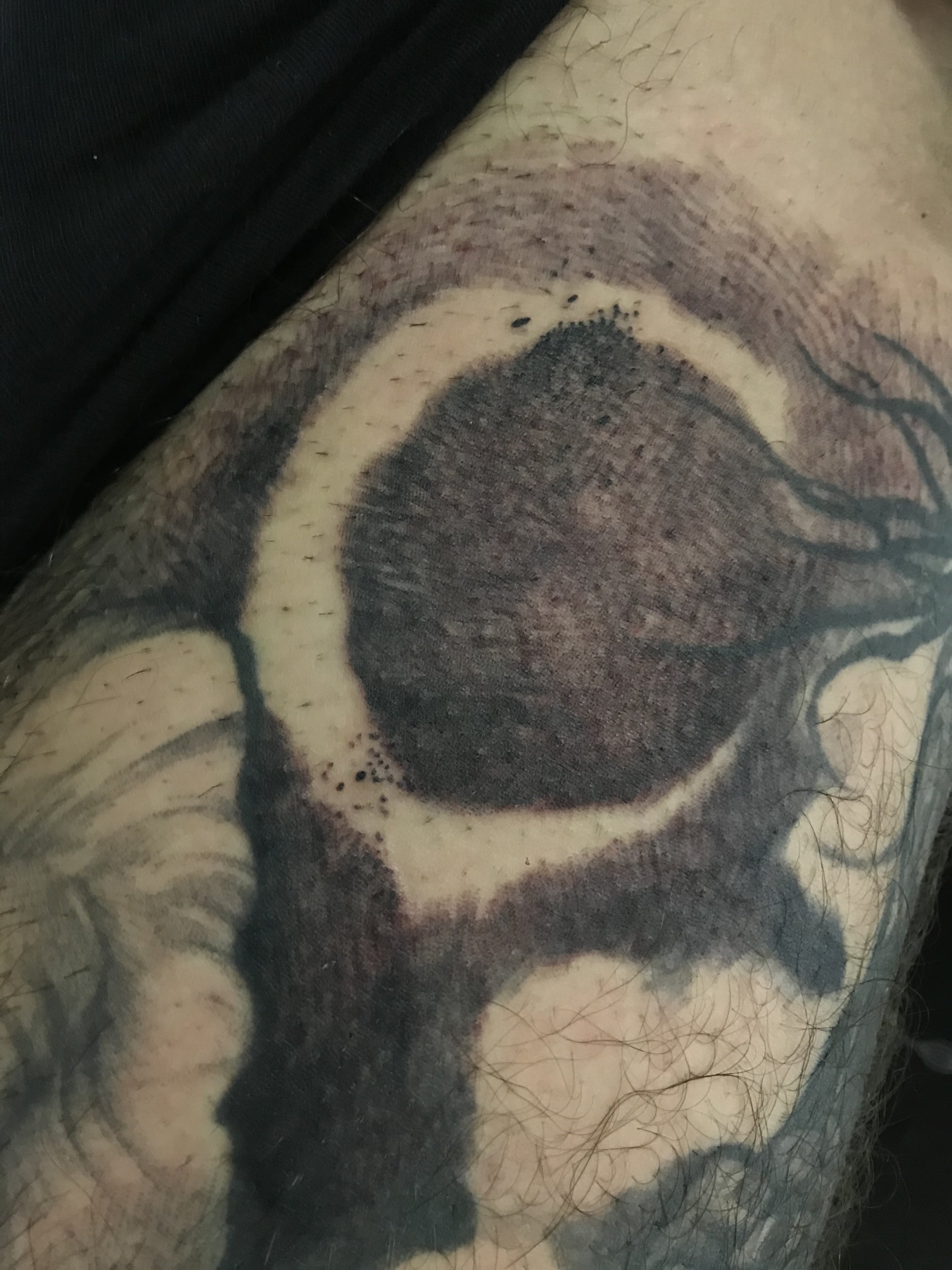 Close-up of a tattoo on a person's arm featuring a detailed circular design with dark shading and lines, surrounded by black ink and some hair.