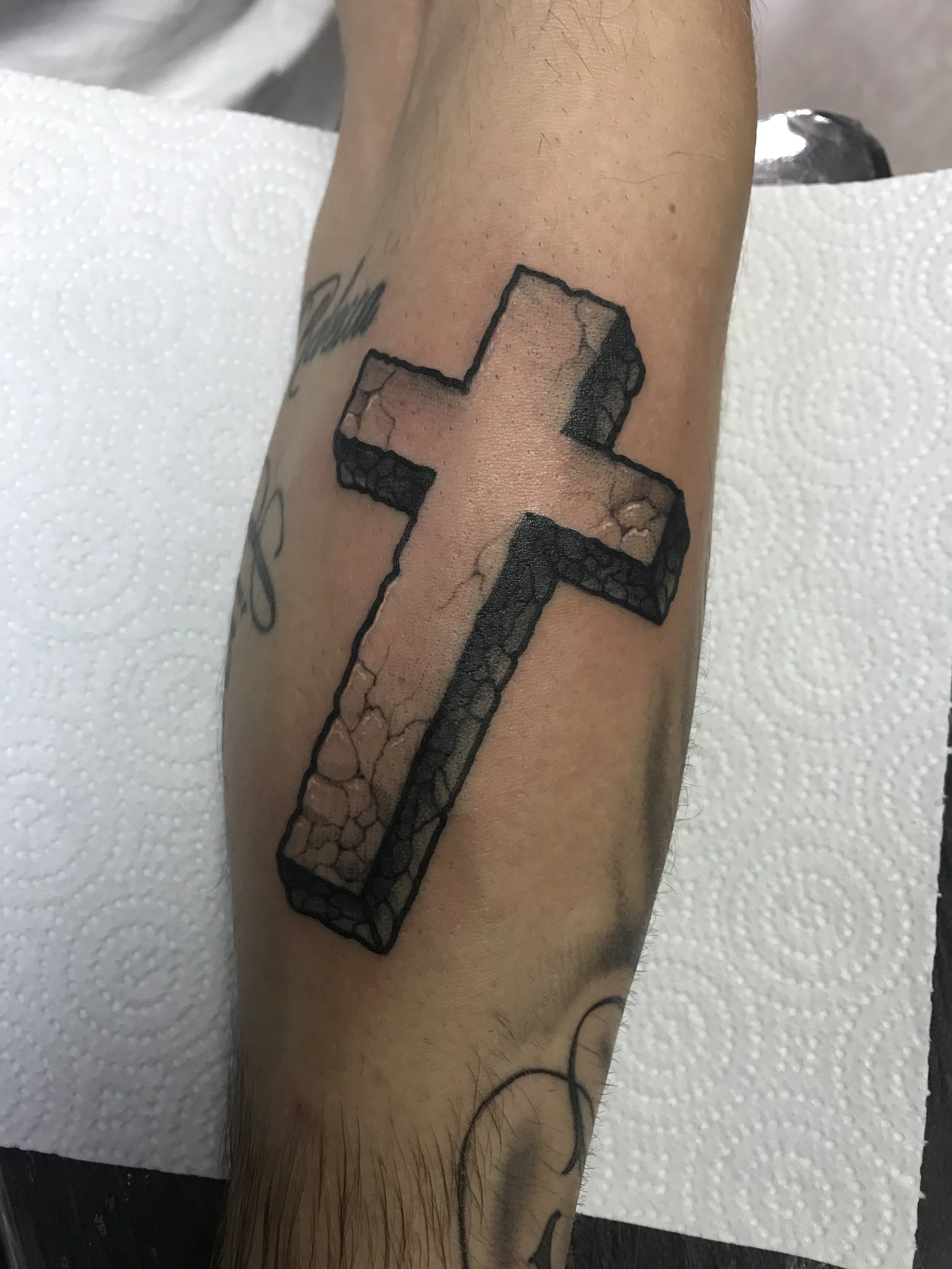 Tattoo of a cross with a textured, stone-like interior on a person's forearm.