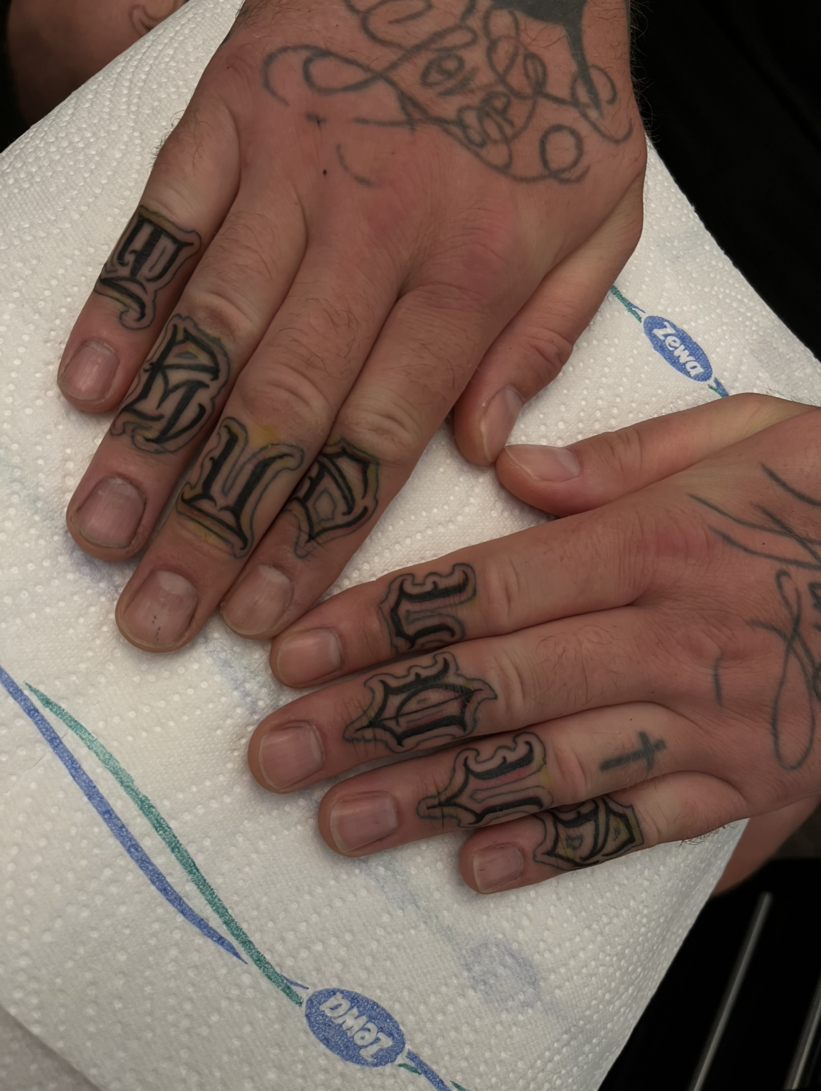 Close-up of a person's hands with numerous black and yellow outlined tattoos, including words and designs on fingers and hands, resting on a white paper towel.