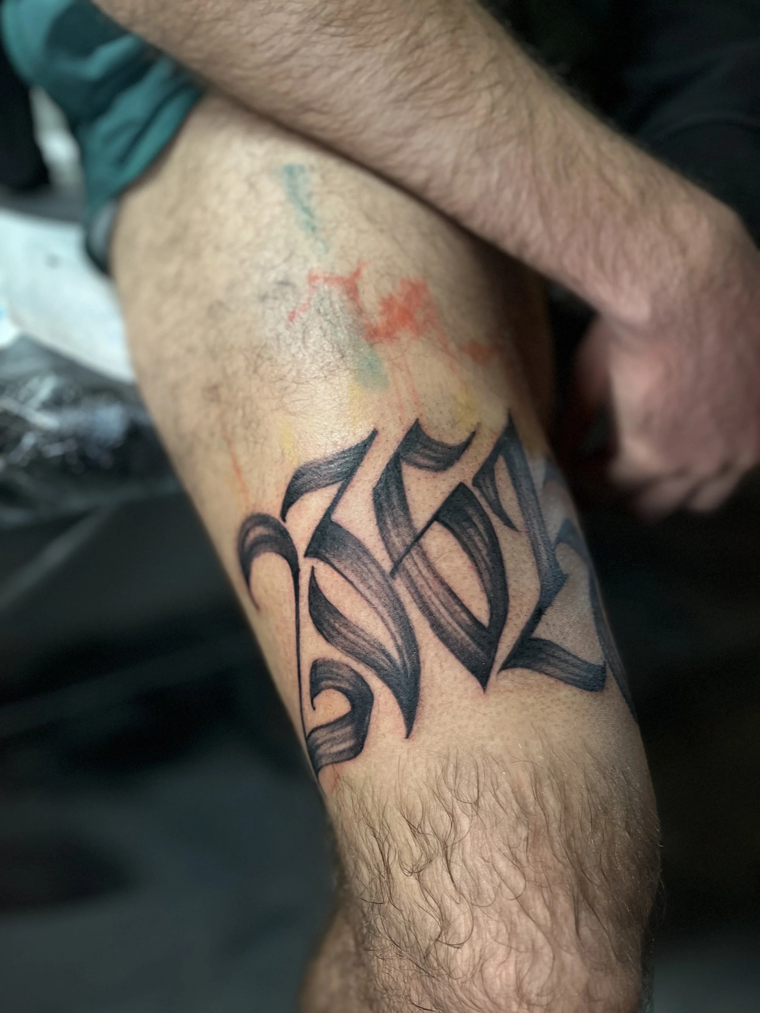 A tattoo of the word 'Koz' in black Gothic-style font on a person's thigh, with colorful watercolor-style splashes above it.