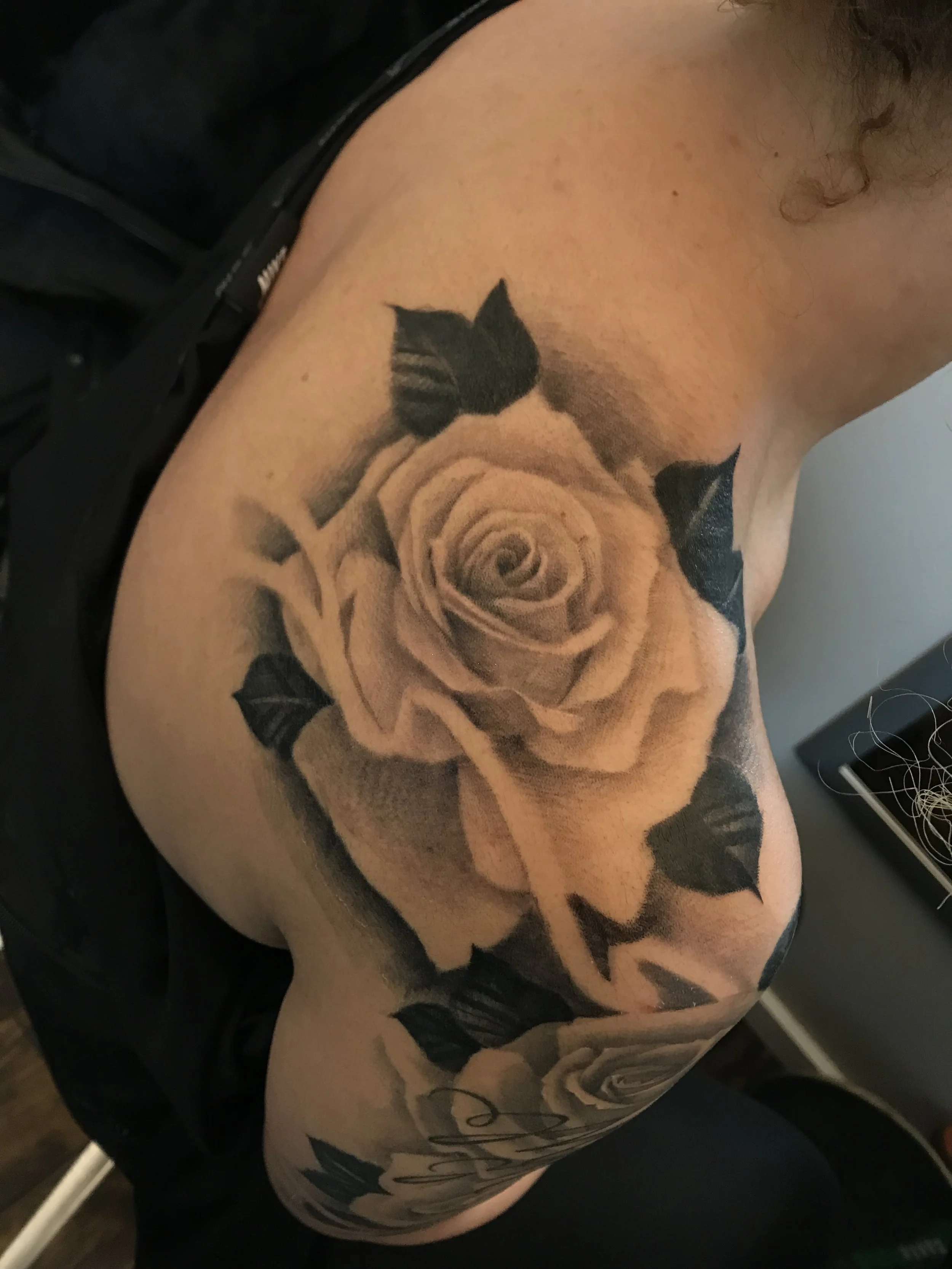 Black and gray shaded tattoo of a rose with leaves on a person's shoulder and upper arm.