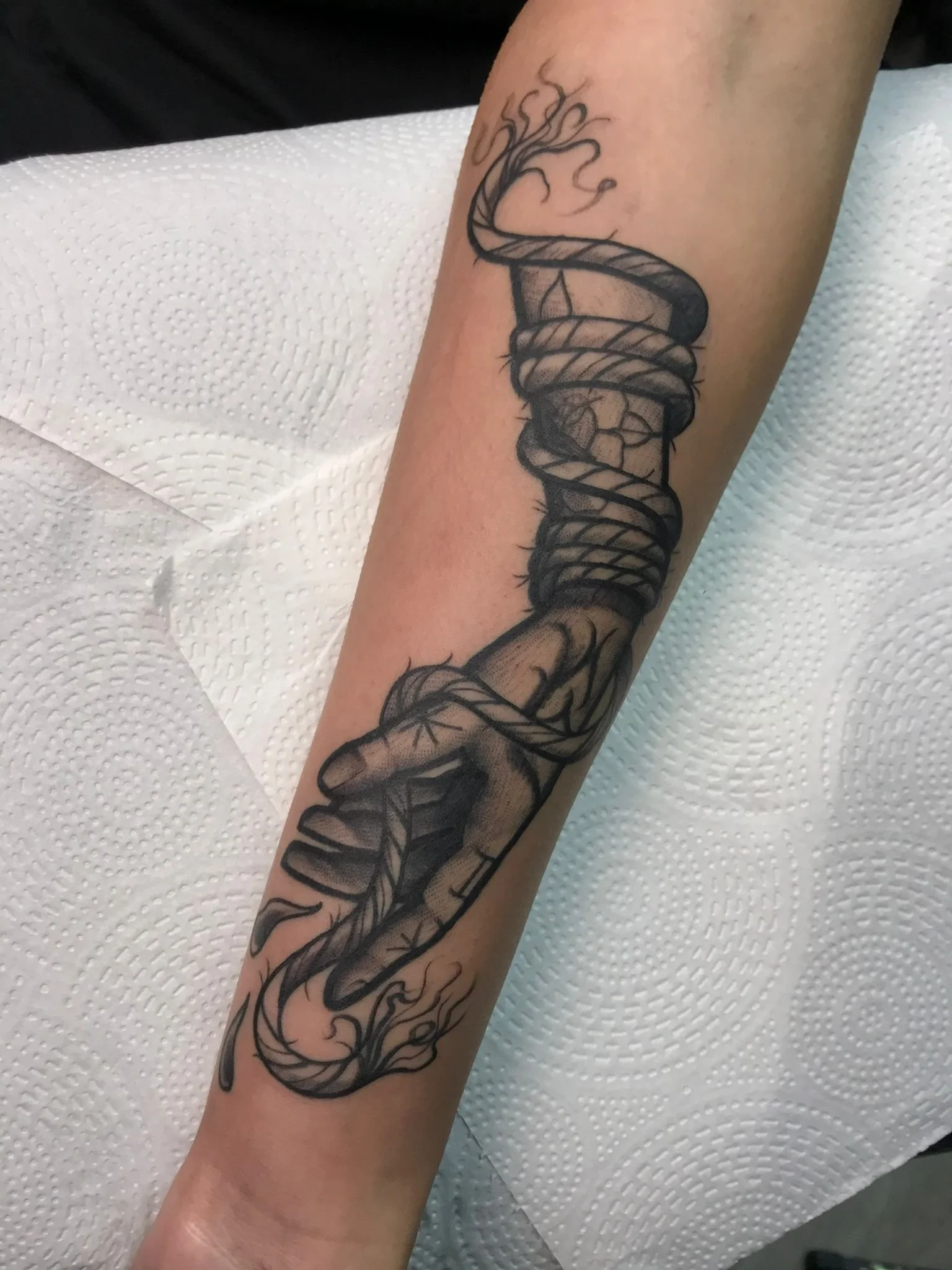 Tattoo of a hand gripping a rope that wraps around a smoking pipe. The tattoo features bold black outlines and shading, with smoke emanating from the pipe.