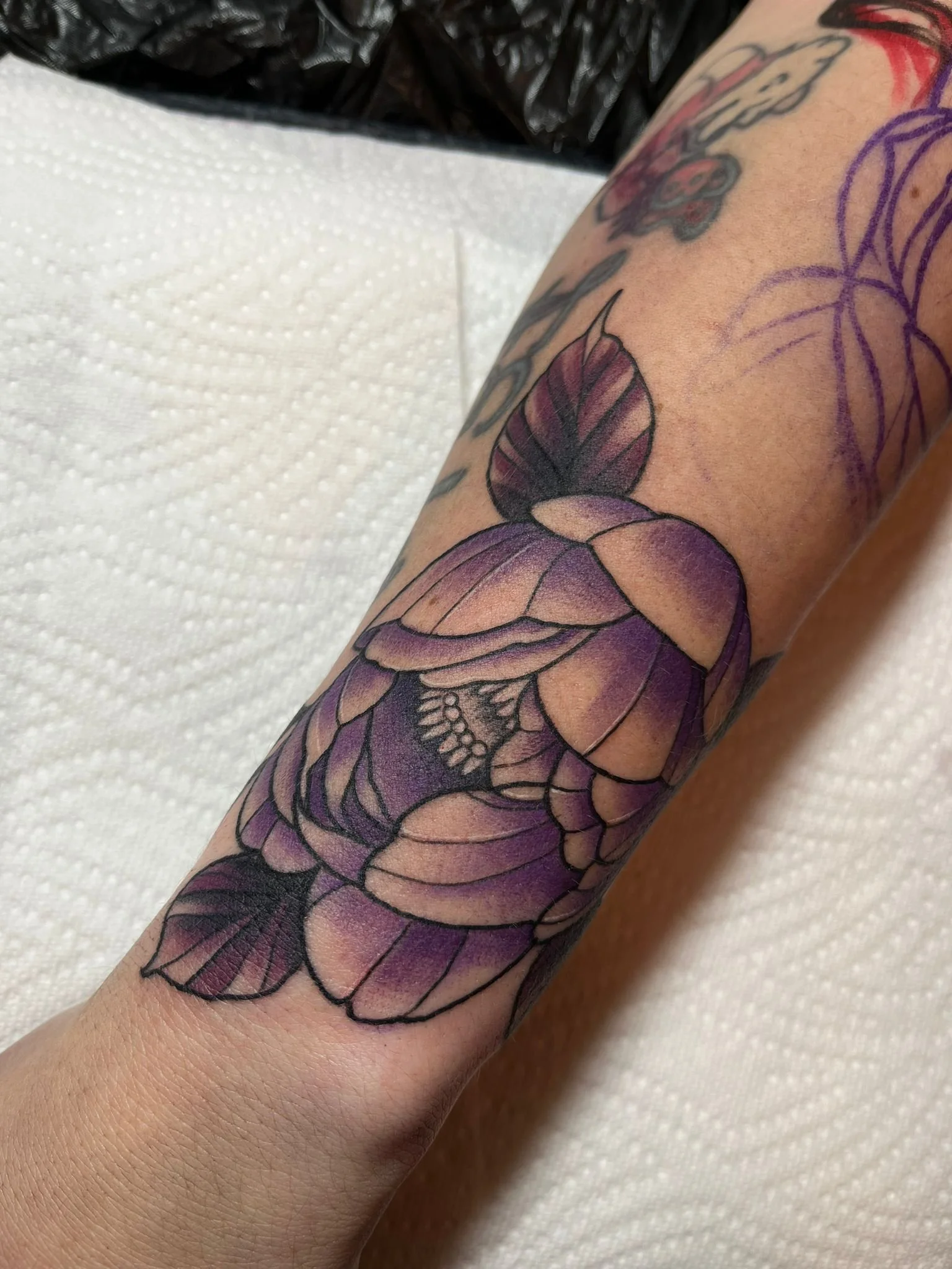 A close-up of a forearm tattoo featuring a large purple flower with green leaves and intricate black line details.