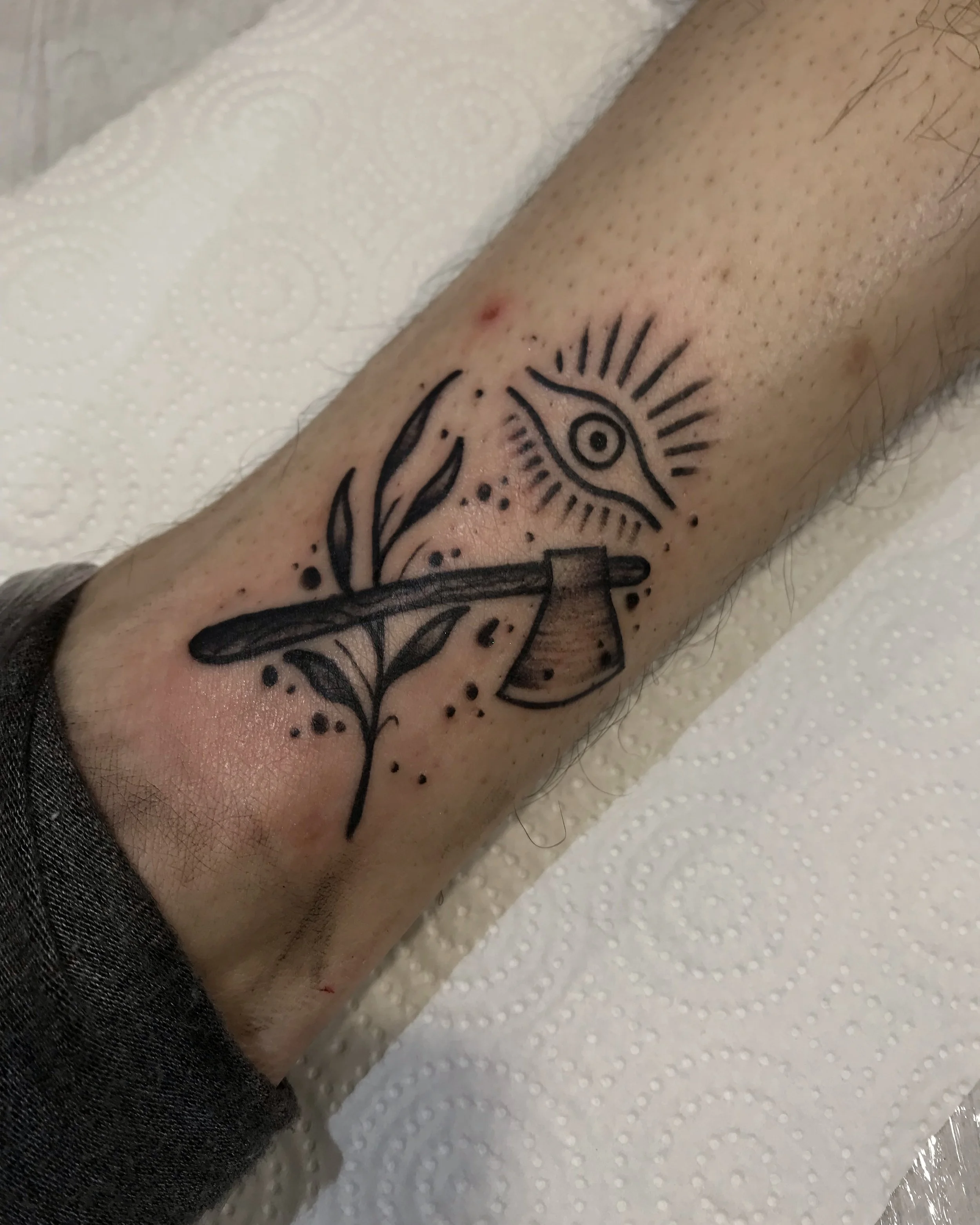 Tattoo on a person's forearm depicting an eye with radiating lines above an axe with leaves, with abstract ink splatters.