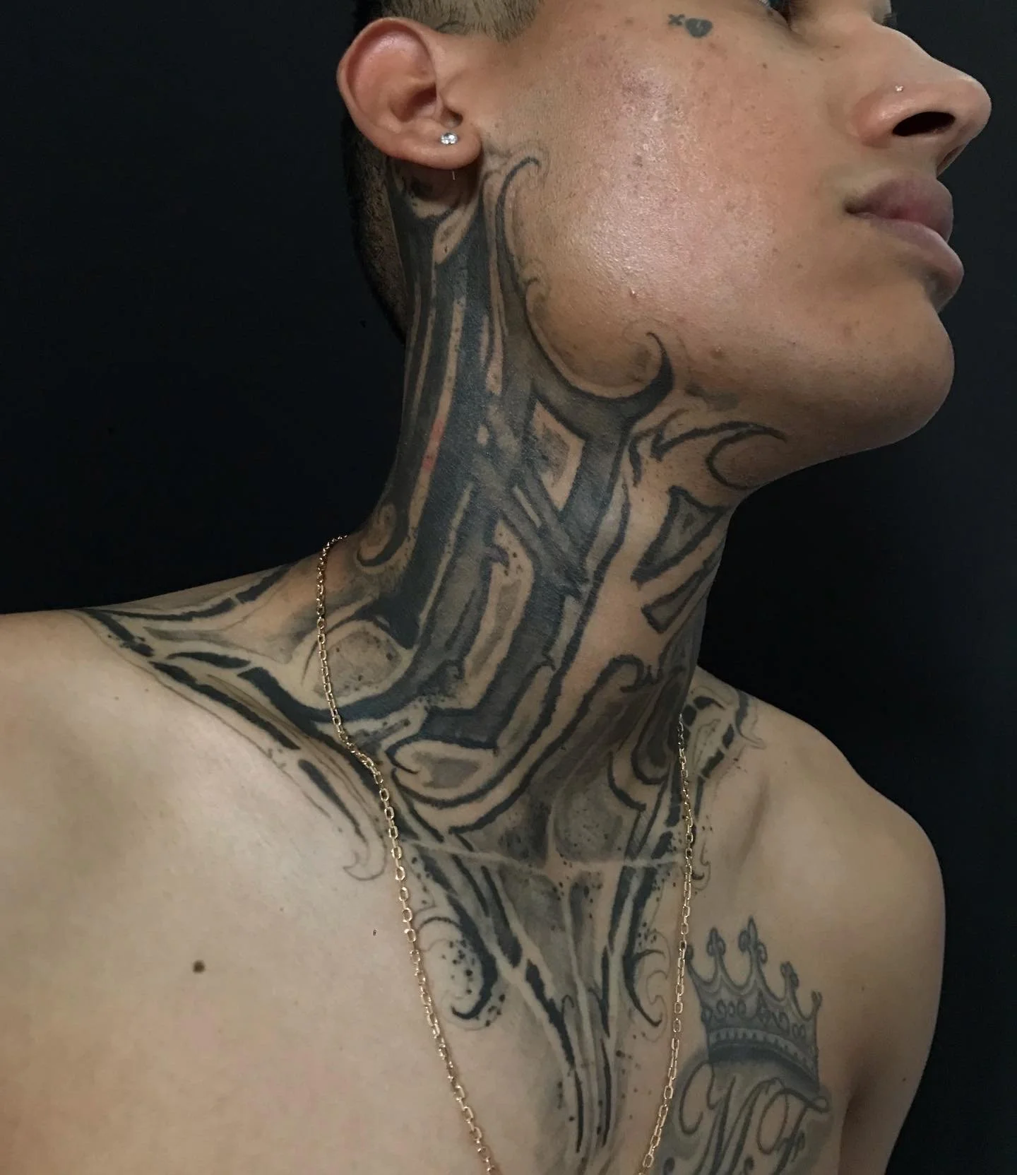 Close-up of a person with a large neck tattoo, earrings, and a chain necklace, with a black background.