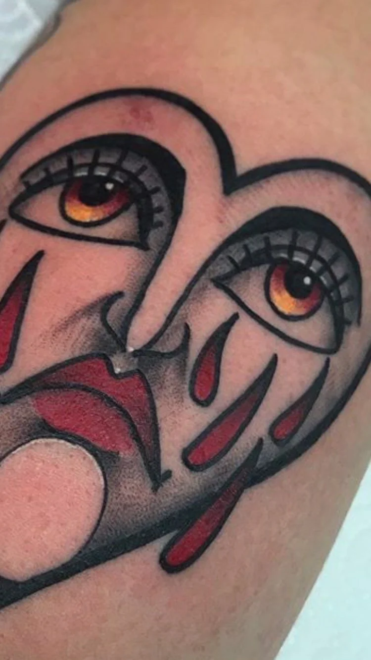 Tattoo of a stylized face with exaggerated features, including large eyes with orange and yellow irises, red pupils, long black eyelashes, and a face with red markings and black outlines.