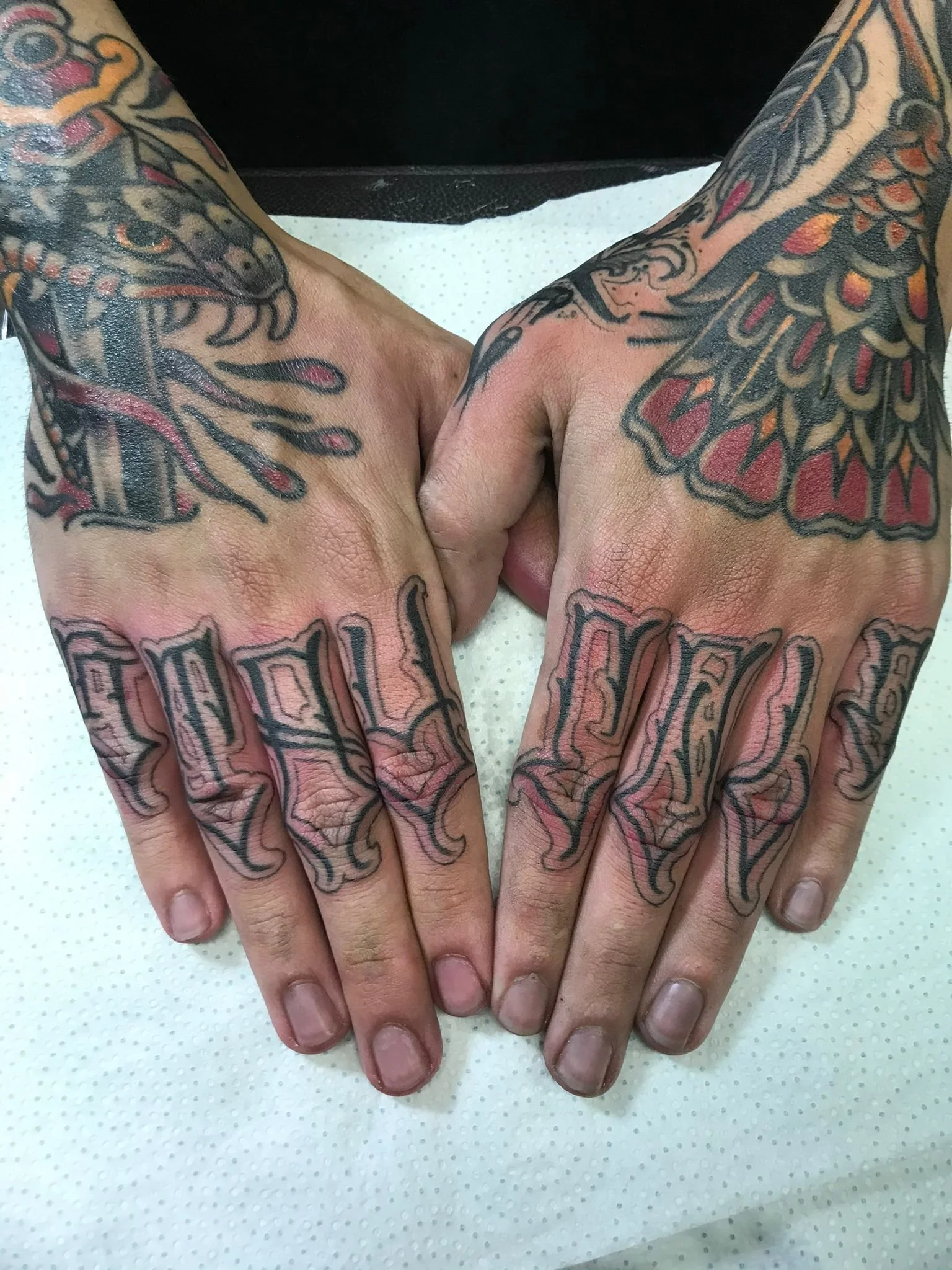 Close-up of two tattooed hands with fingers touching, resting on a white surface. The tattoos include colorful designs on the backs of the hands and fingers with lettering on the fingers.