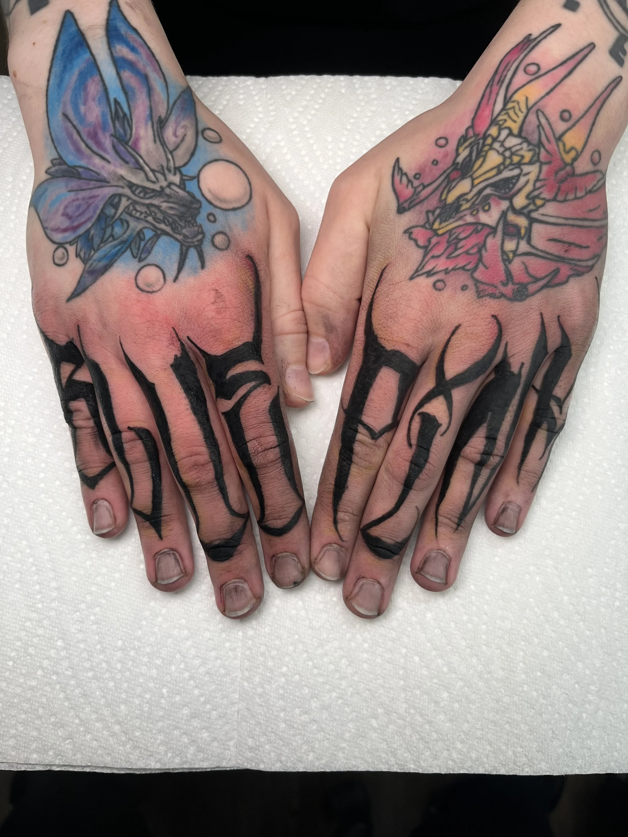 Tattooed hands with bold black text across fingers, featuring a blue dragonfly on the left hand and a pink and yellow dragon on the right hand, both on a white surface.