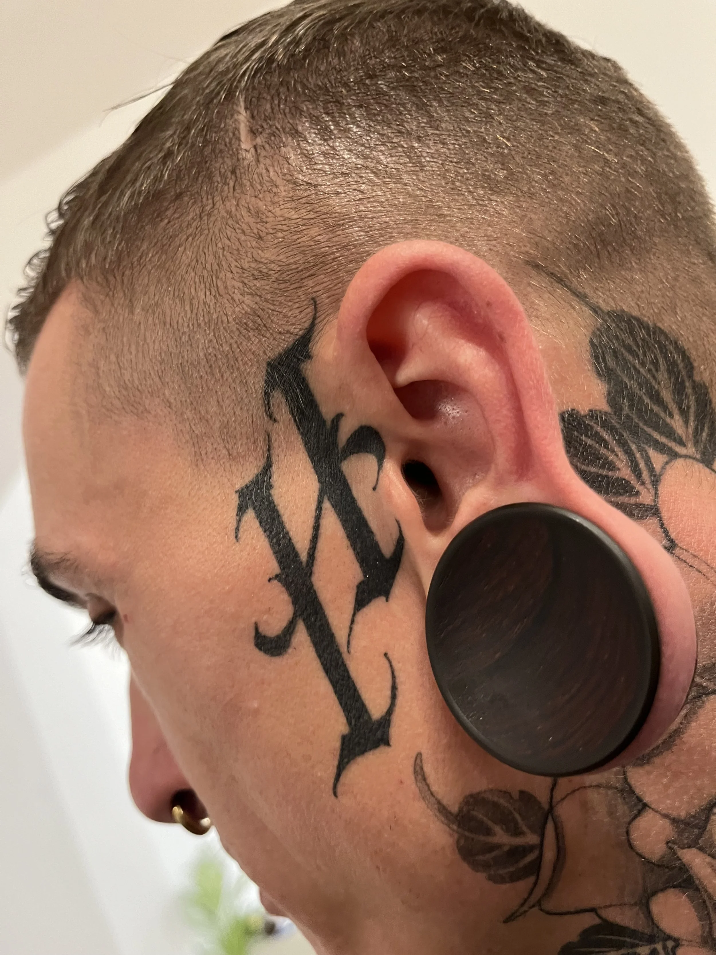 Close-up of a person's face with trimmed hair, earrings, and tattoos, including a large black letter 'N' on the cheek, a black leaf design behind the ear, and a dark spiral ear gauge in the ear.