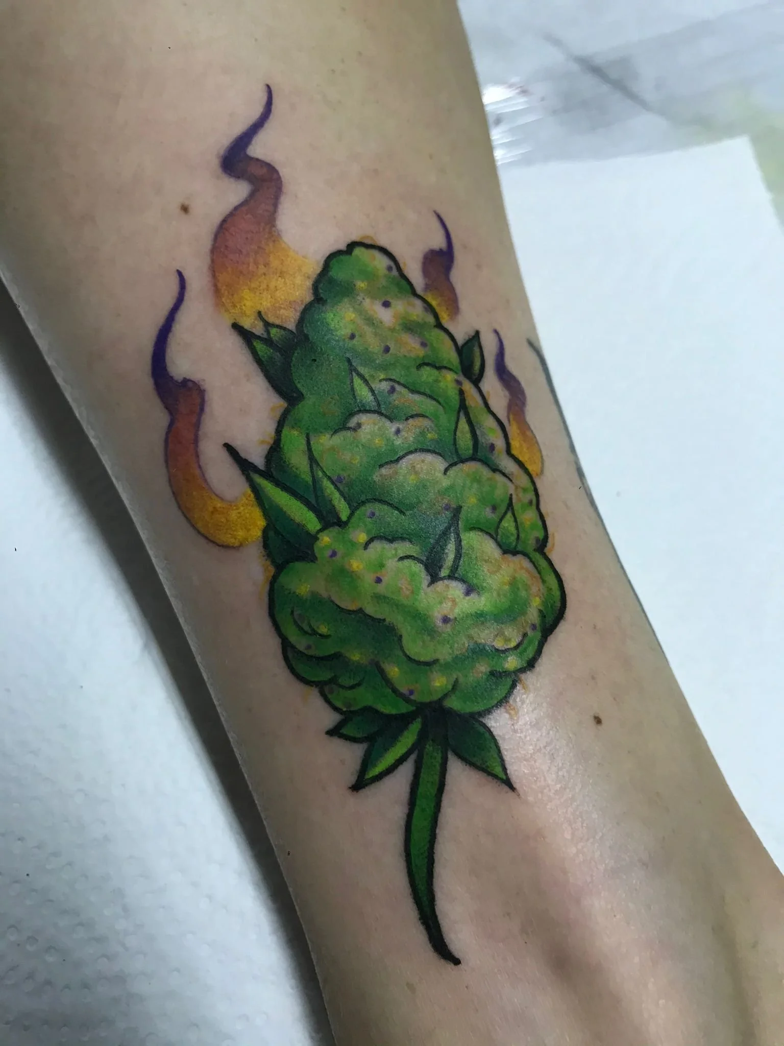 Colorful tattoo of a green cannabis bud with purple and yellow flames in the background on a person's arm.