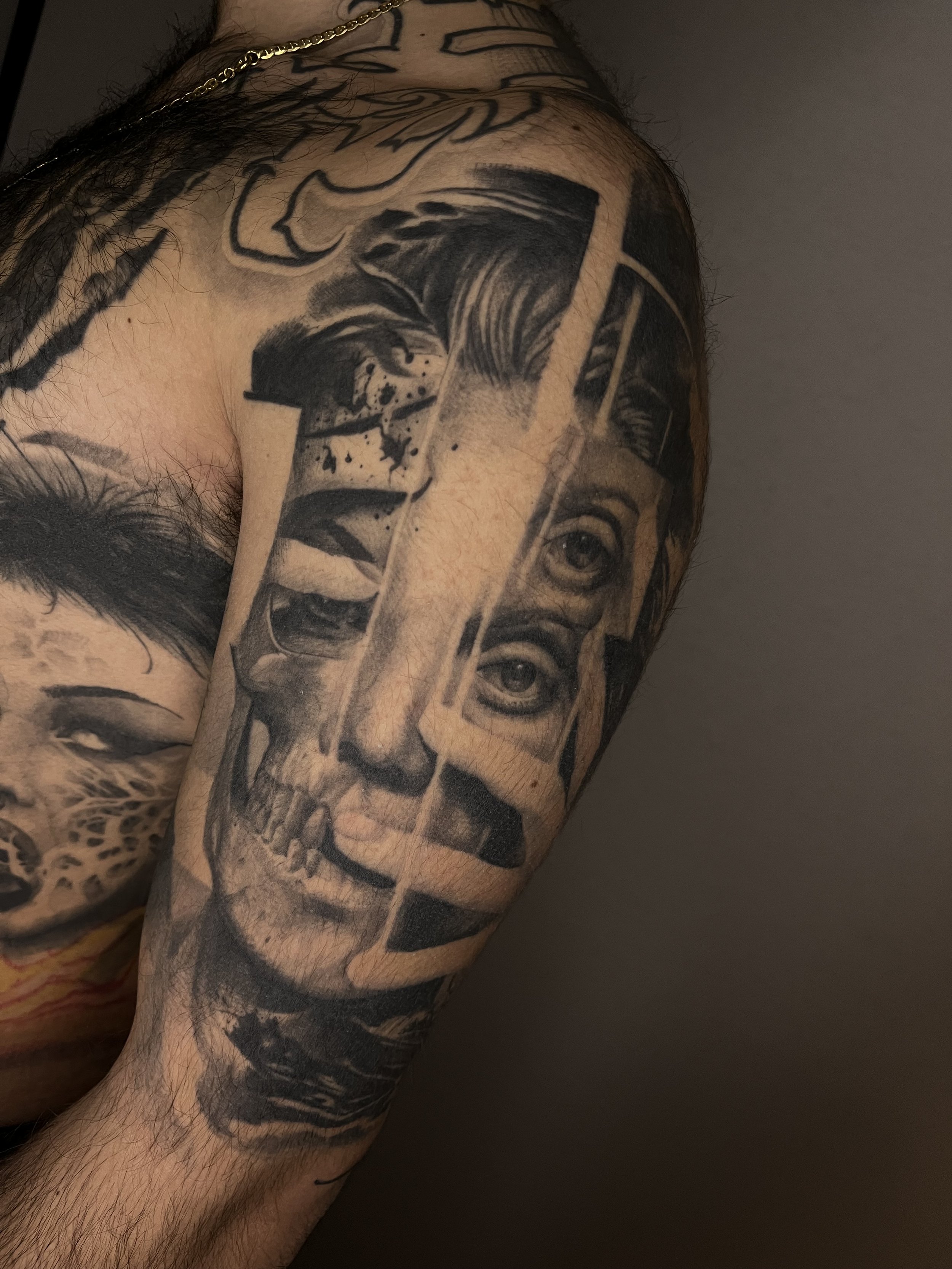 Close-up of a person's upper arm and shoulder tattoos, including a realistic portrait of a woman with two eyes and a smiling mouth, and other abstract designs in black and gray ink.