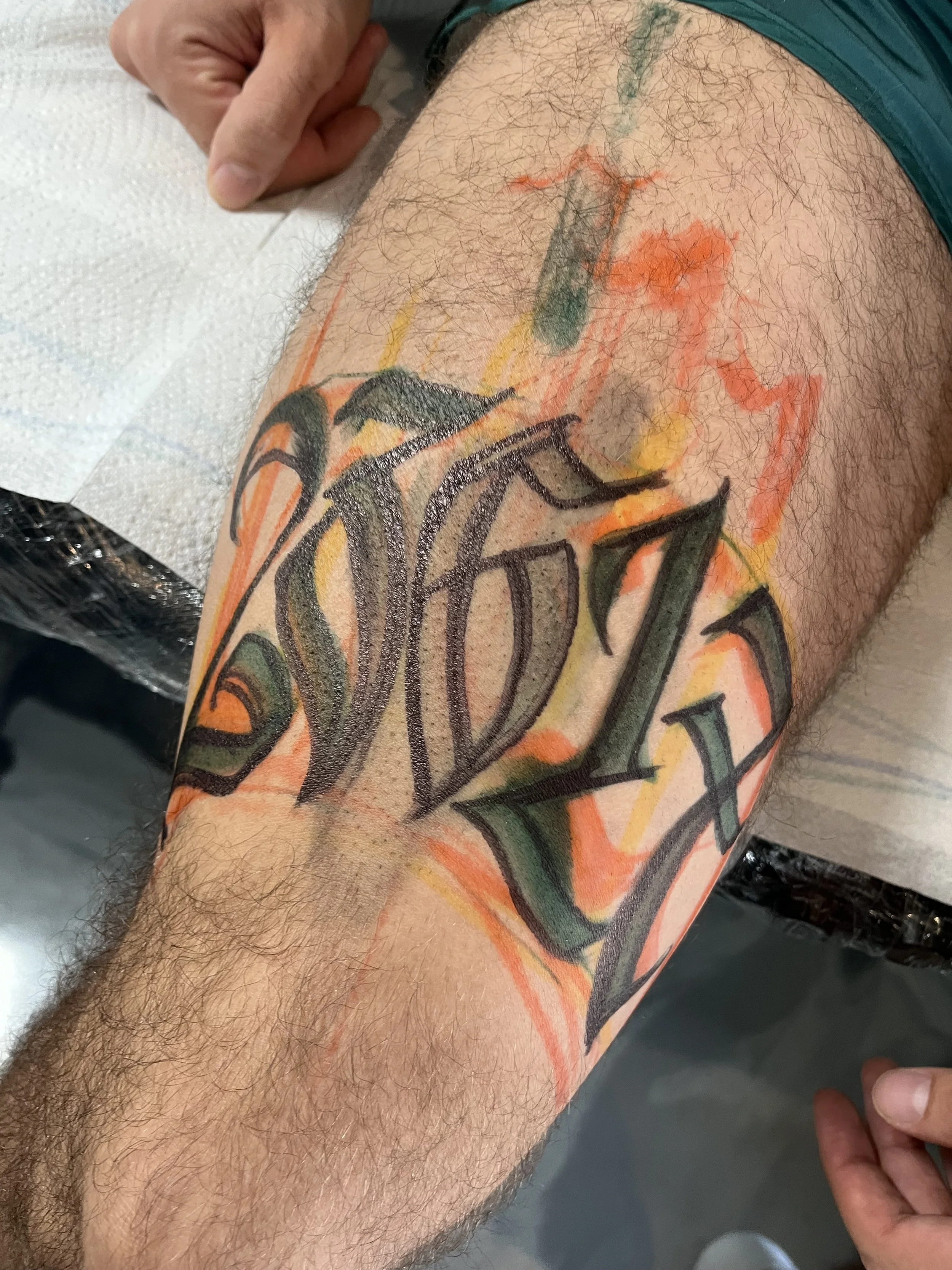 A person's thigh with a tattoo of stylized black lettering, with some orange, yellow, and green shading, and a green sketch outline nearby, hinting at a work-in-progress tattoo.