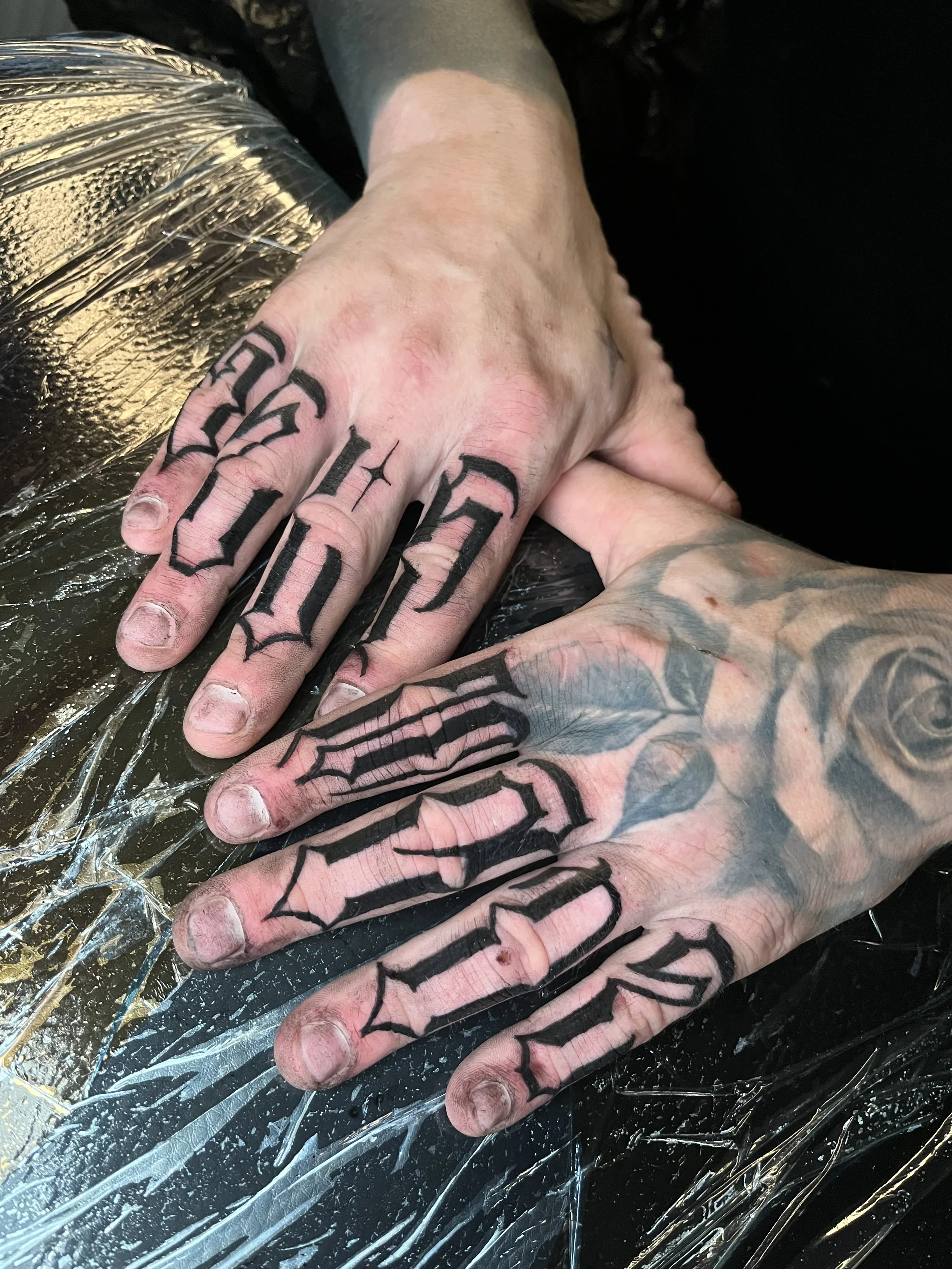 Two hands with black gothic-style tattooed lettering on the fingers and a large tattoo of roses and leaves on the back of the hand, resting on a black surface covered with clear plastic wrap.