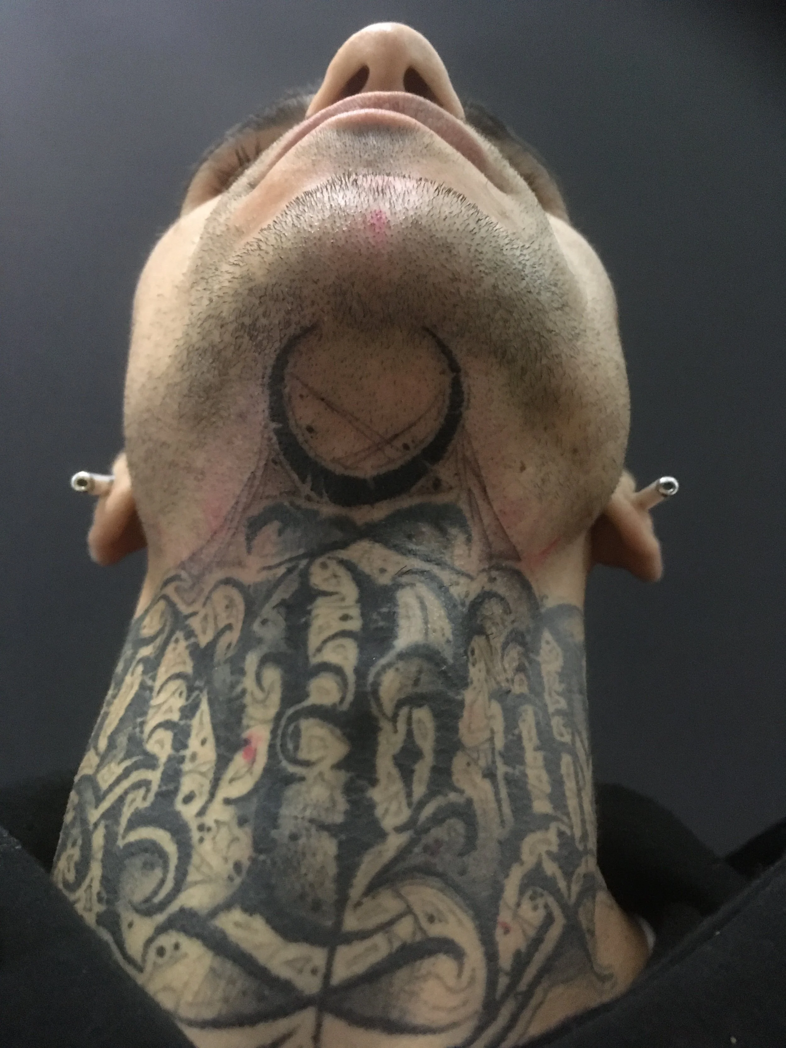 Close-up view of a person's face and neck, showing tattoos and piercings, with an upward camera angle.