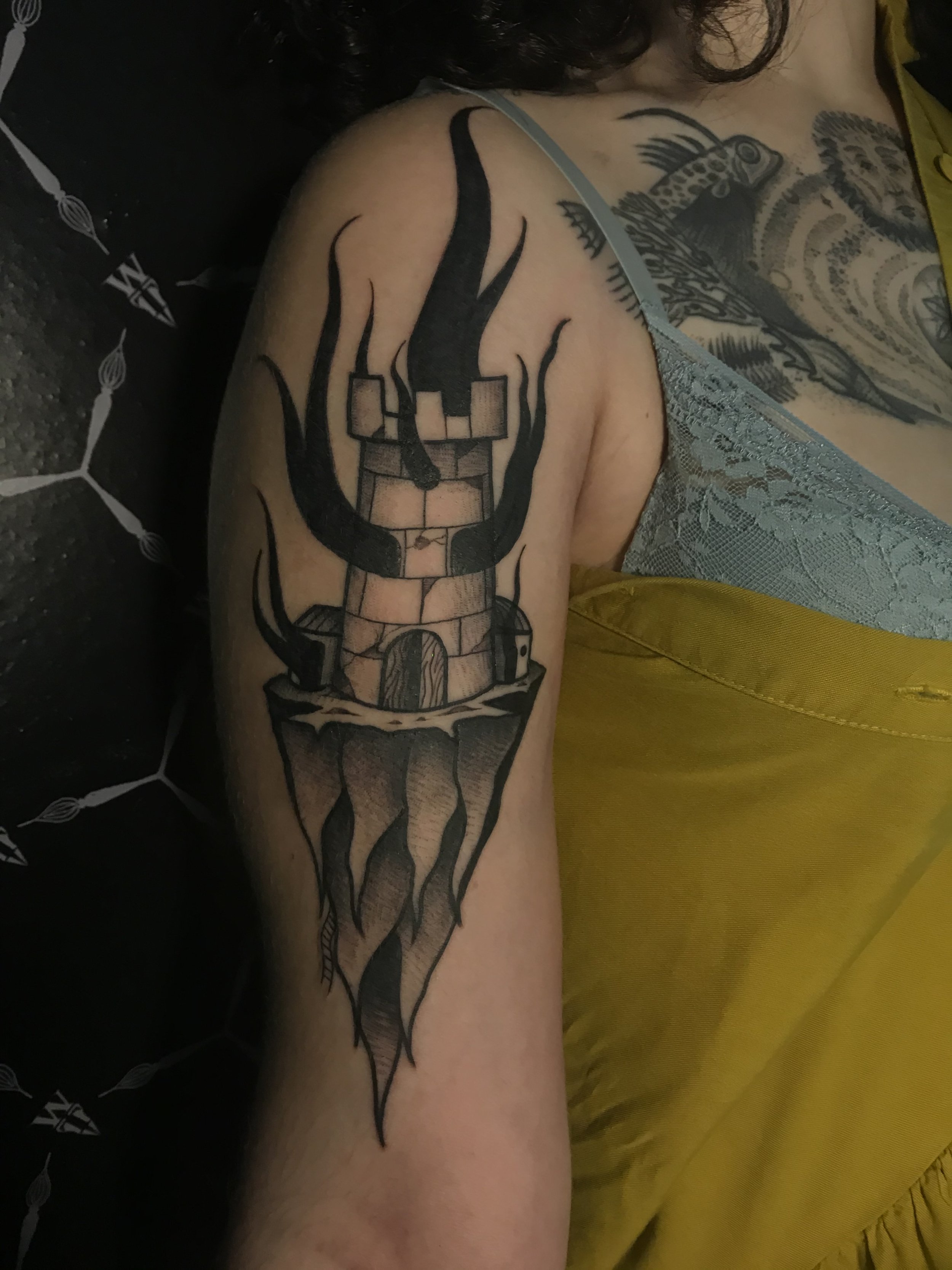 Tattoo of a lit torch with a brick base and flames extending upward, located on the person's upper arm.