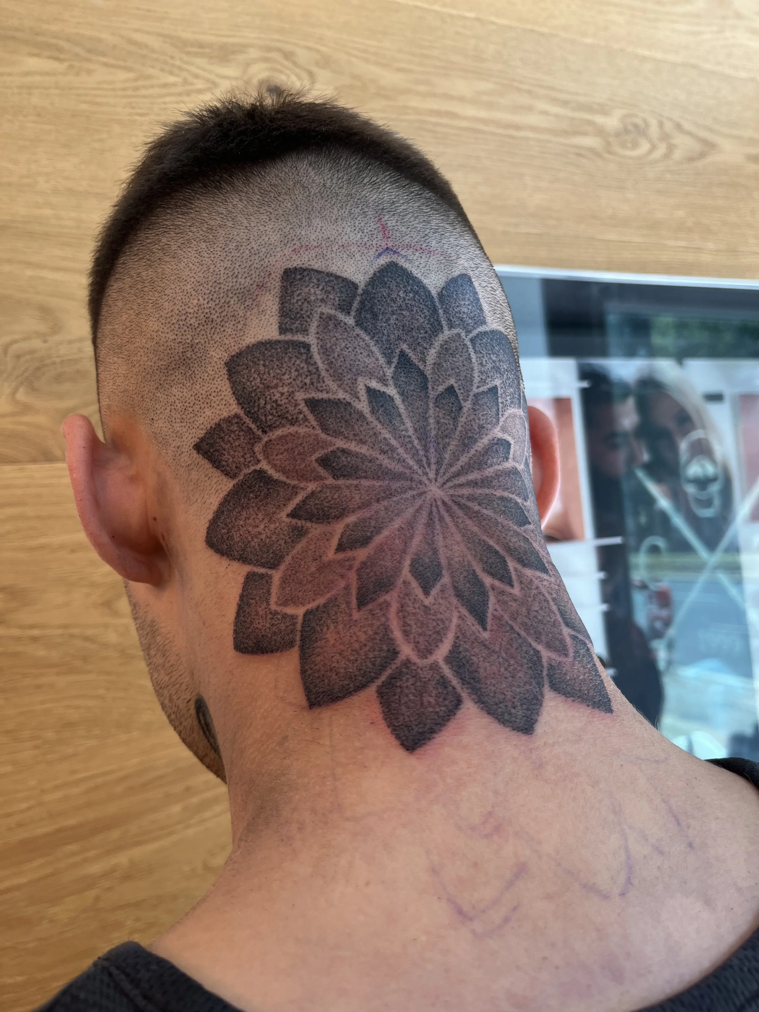 A person's head with a large black tattoo of a floral pattern on the back of the neck, with a second smaller pattern faintly visible below it.