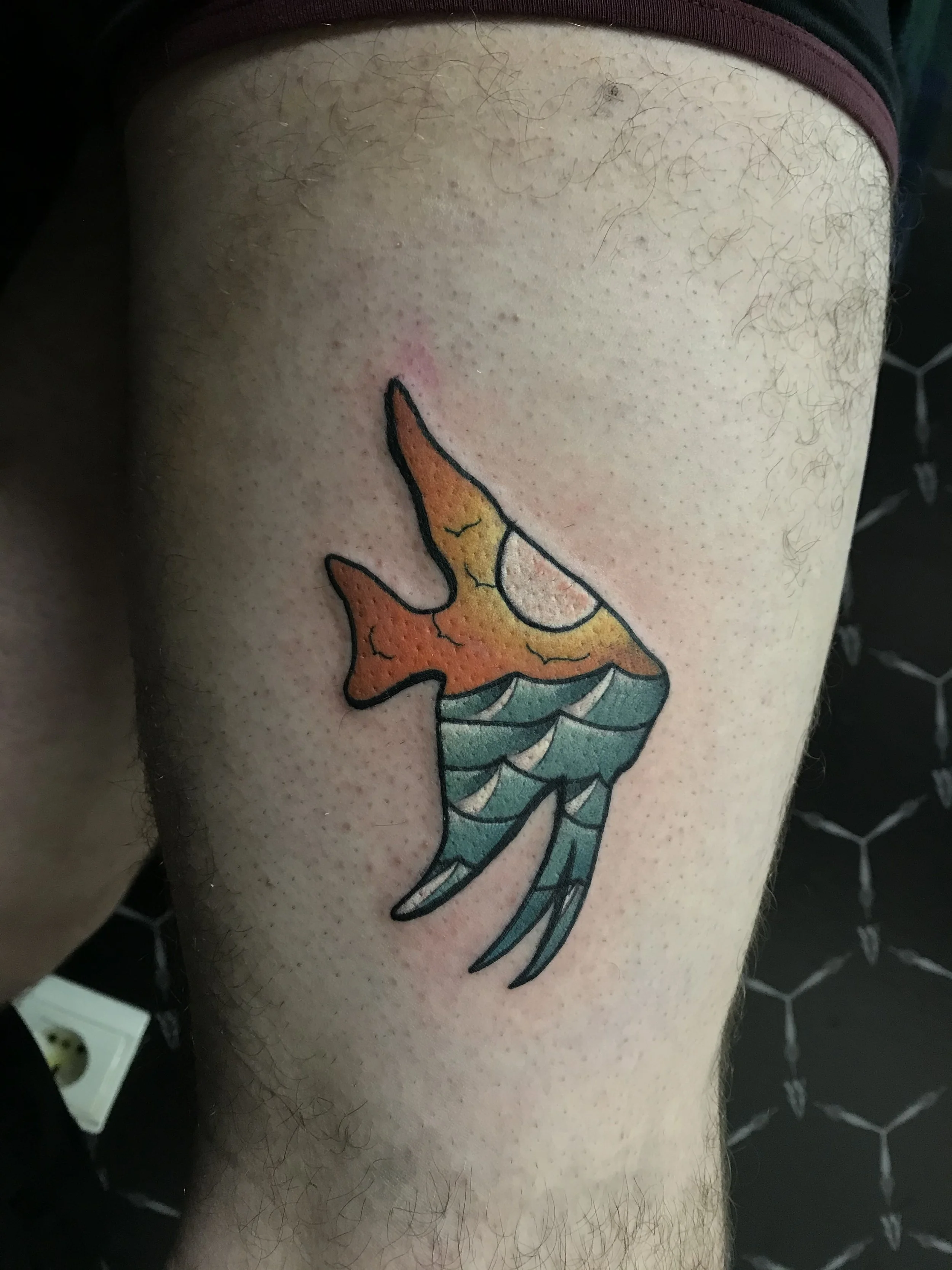 Colorful tattoo of a hummingbird on a person's thigh, with orange, yellow, and turquoise colors, outlined in black.