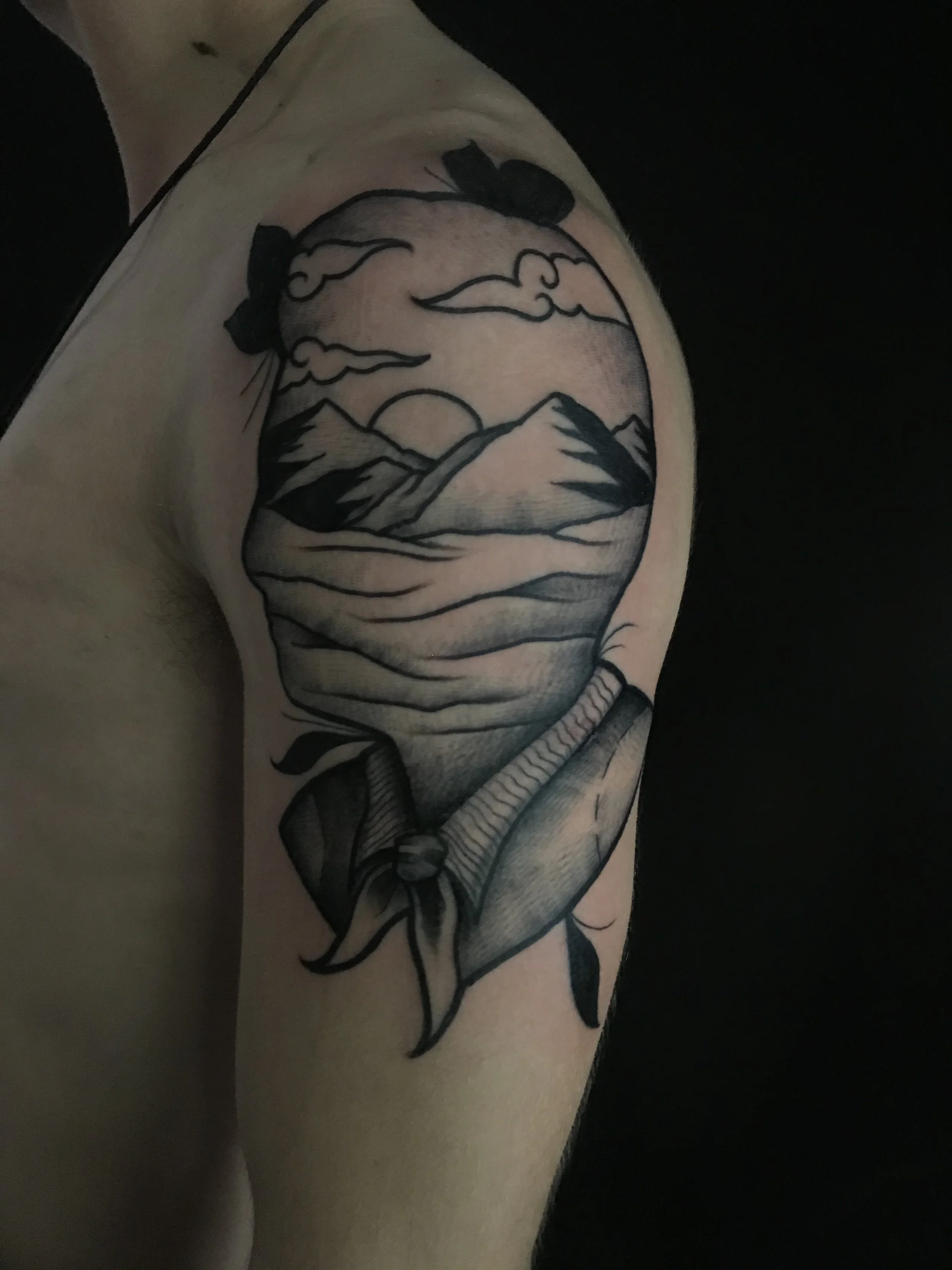 Black and gray tattoo of a landscape with mountains, clouds, and a setting or rising sun, partially covered by a bandana or scarf, on a person's upper arm.