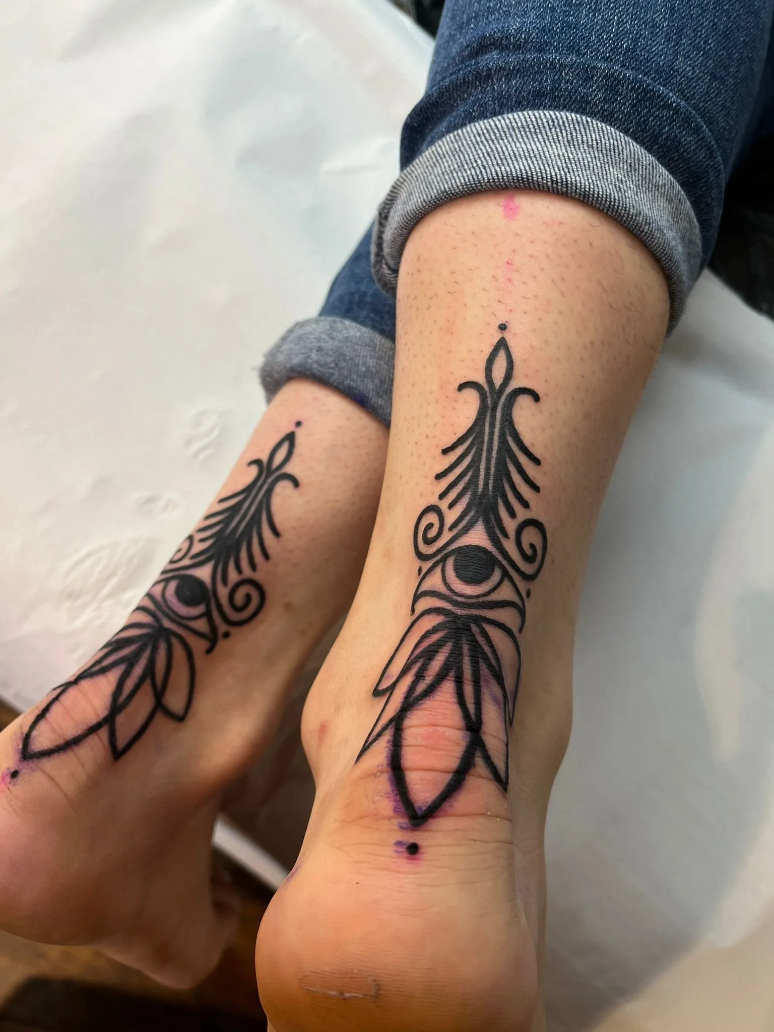 Fresh black tribal-style tattoos on the person's ankles and feet, with their legs crossed and wearing rolled-up jeans.