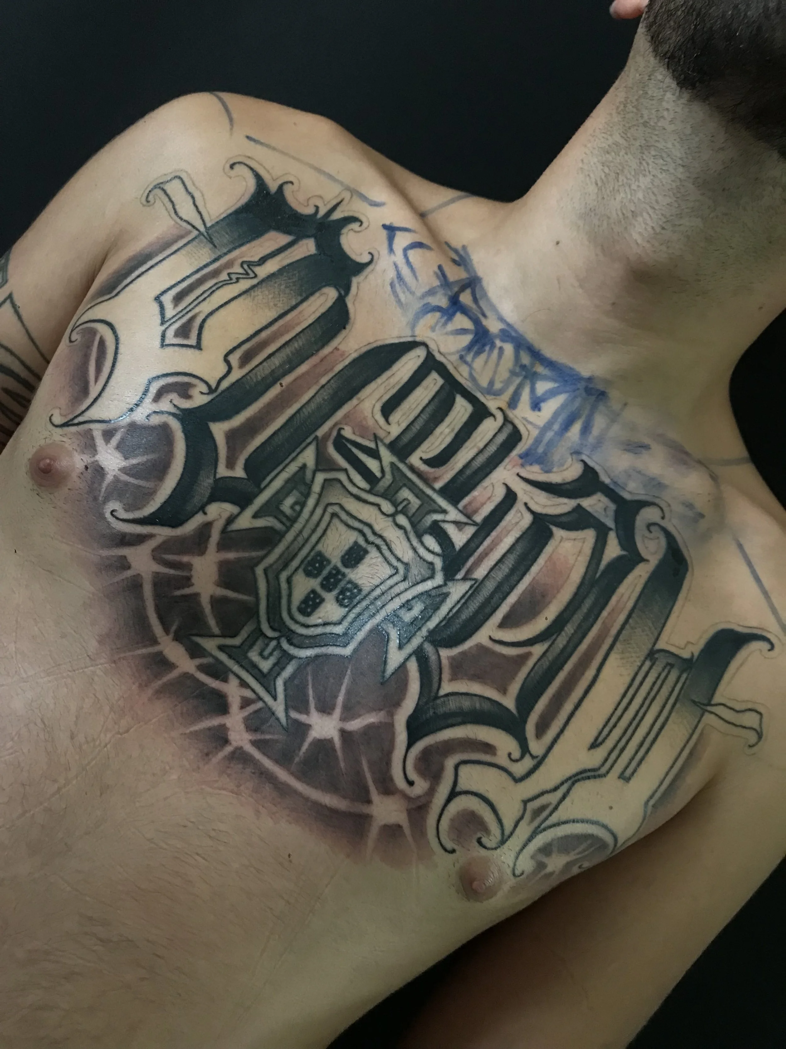 A person's chest with a large, detailed black tattoo of the Portugal coat of arms, shield, and elaborate decorative elements.