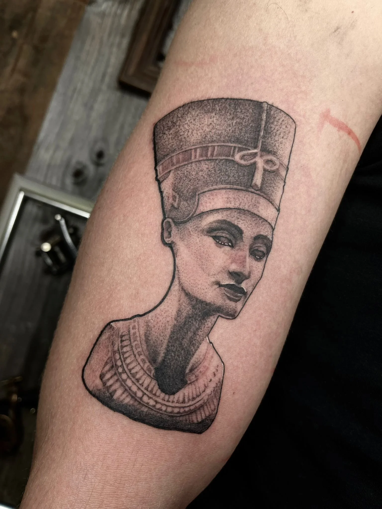 Black and gray tattoo of an Egyptian queen with a tall headdress and detailed clothing, located on a person's forearm.