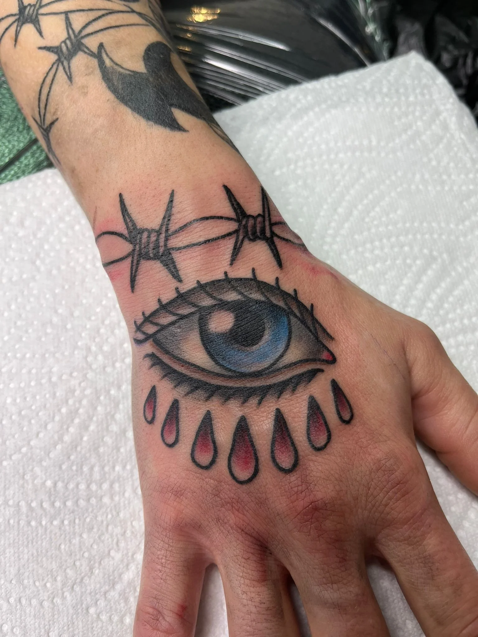Tattoo of an eye with tears on the hand, barbed wire above the eye, and part of a bird with a body and beak visible on the wrist.