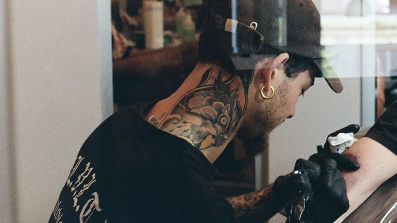 Tattoo artist with multiple tattoos, wearing a black cap and gloves, in the process of tattooing a person's arm in a tattoo studio.