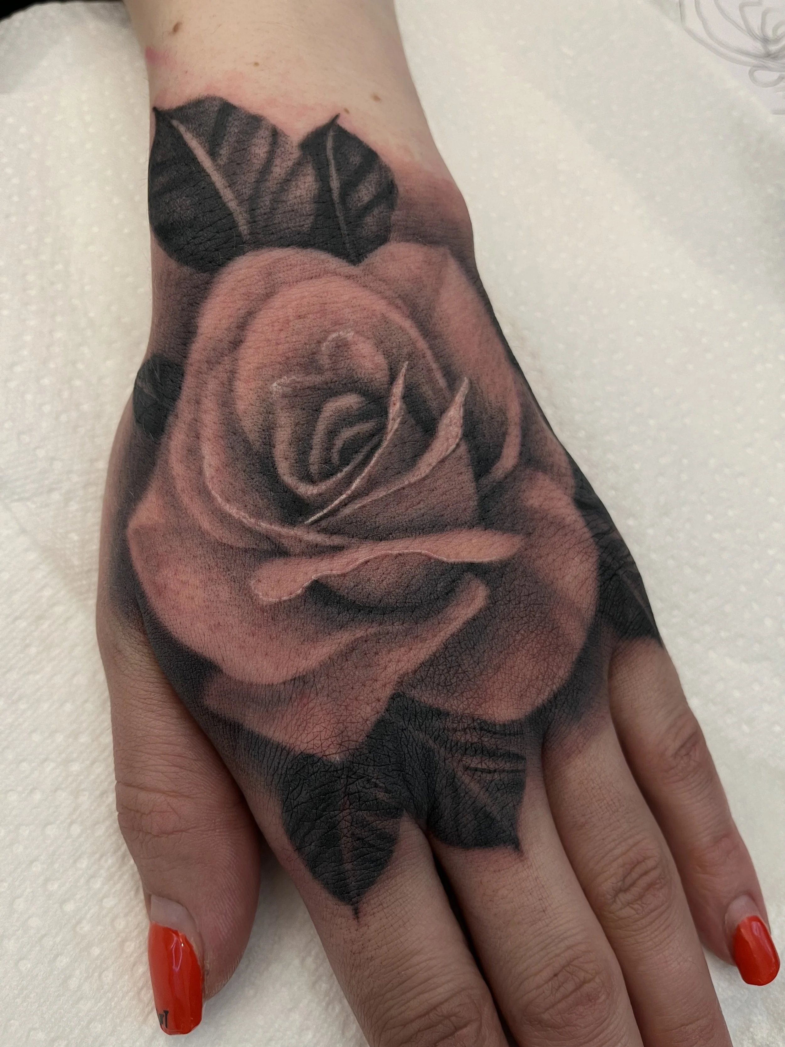 A realistic black and gray tattoo of a rose on the top of a person's hand and wrist.