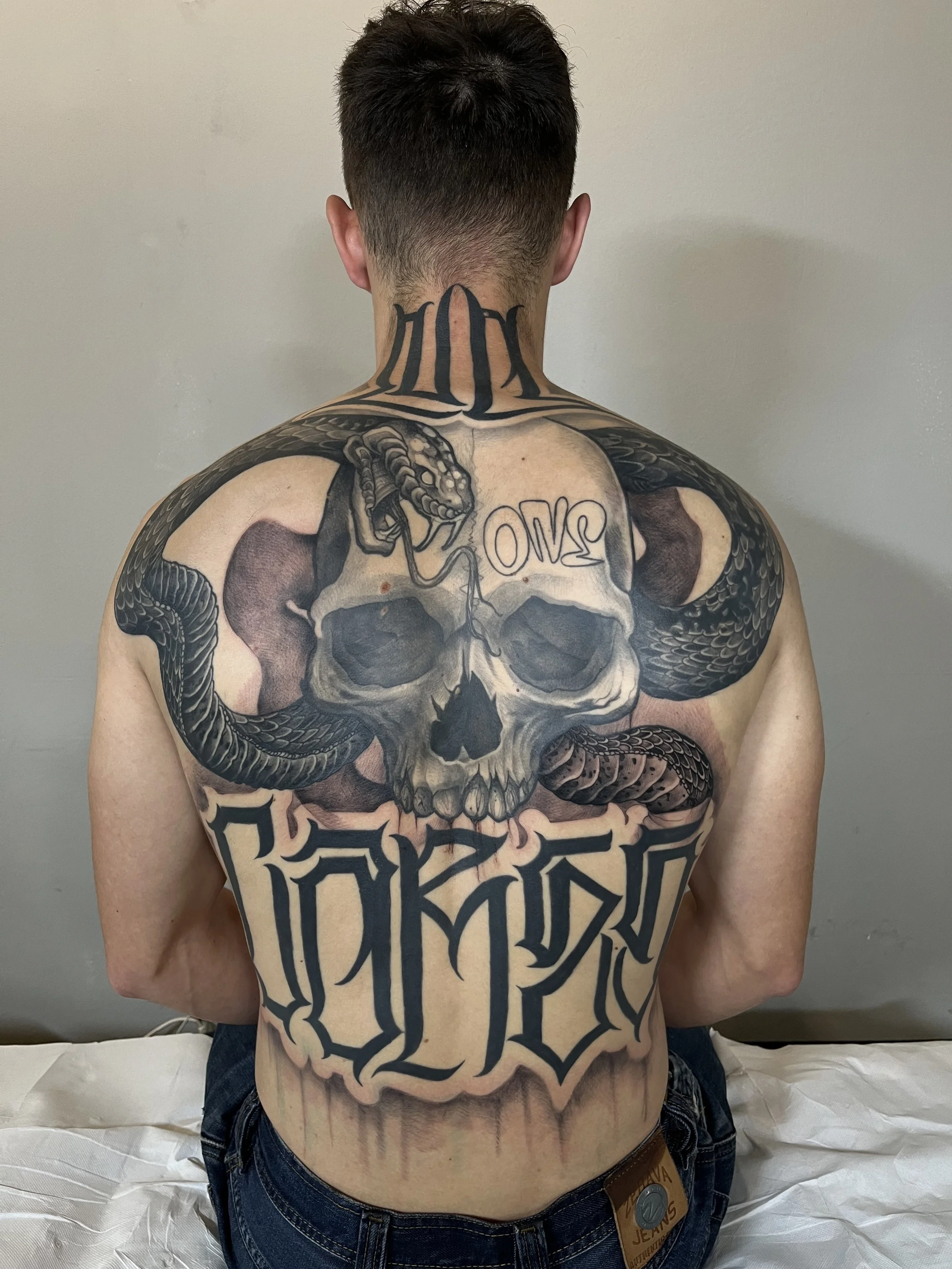 Back view of a man with extensive tattoos, including a skull with snakes and the word 'OMN' on the skull, with large gothic script lettering across the lower back.
