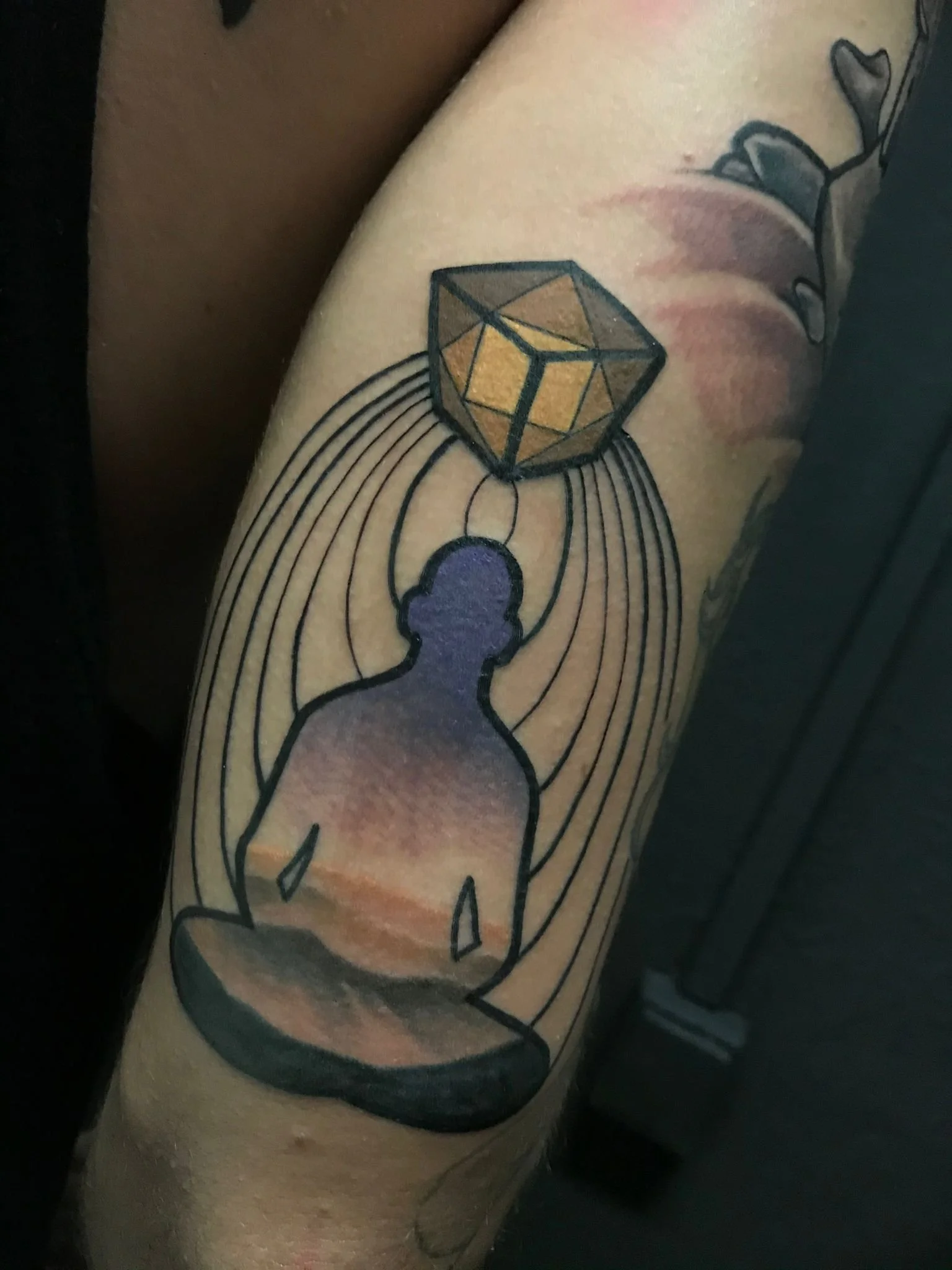 Tattoo of a human figure seated in meditation, with a geometric gemstone above the head, surrounded by radiating lines, and a background resembling a sunset.