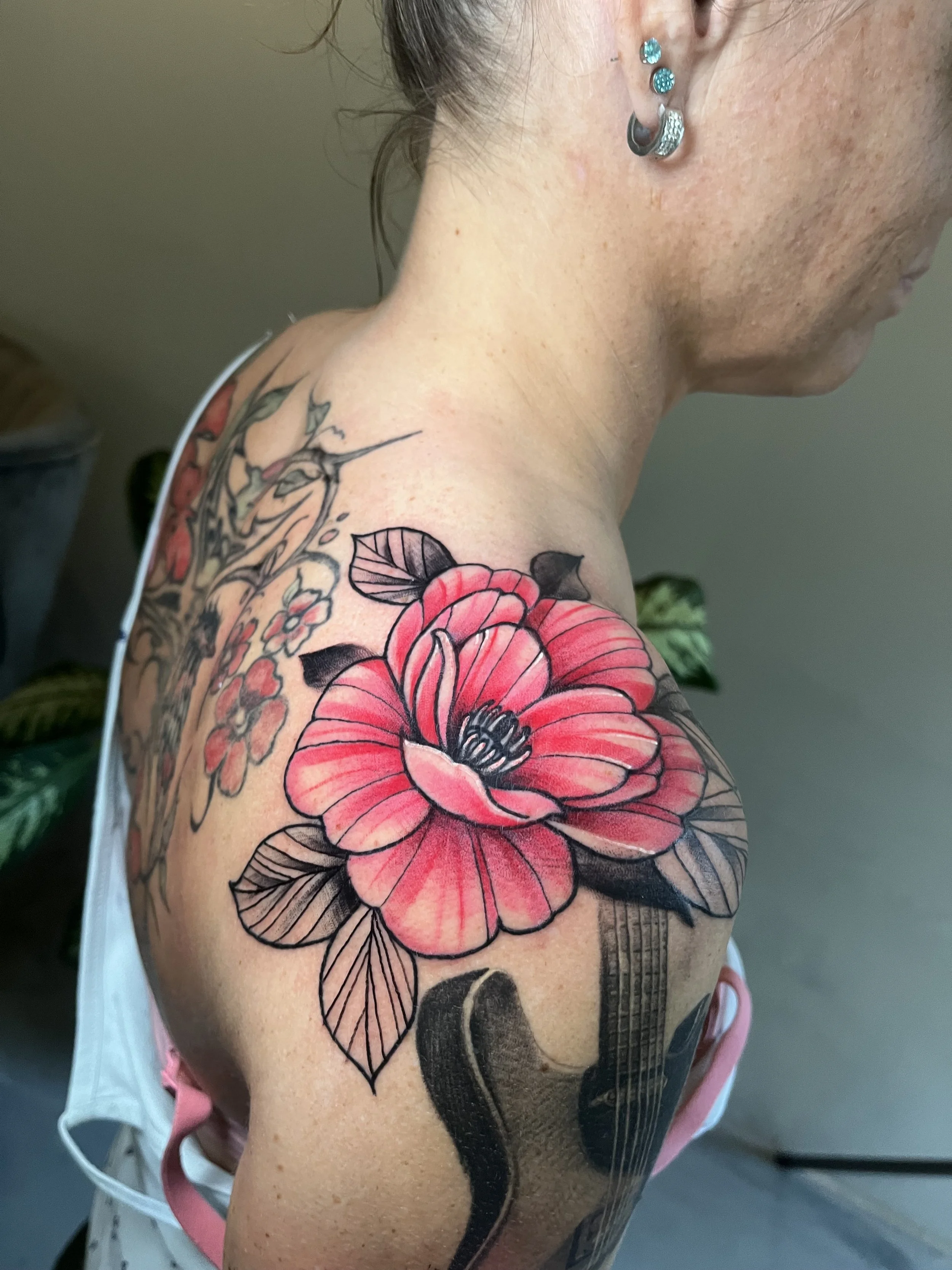 A woman with multiple tattoos on her shoulder and neck, including a large pink and black flower tattoo on her upper arm and smaller black and gray floral tattoos on her shoulder. She also has multiple earrings in her ear.